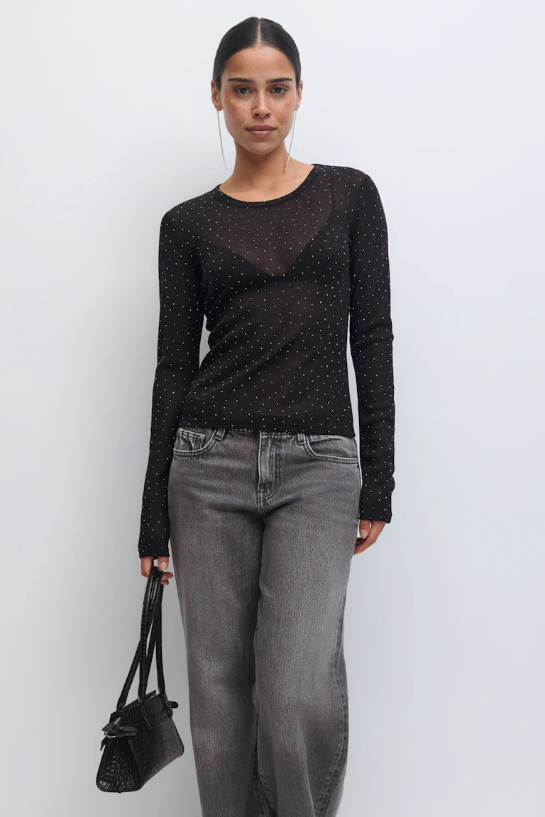 Sheer black long-sleeve top with small metallic dots layered over a black triangle bralette, faded grey high‑waist straight-leg jeans, small black croc‑embossed handbag, and long drop earrings.