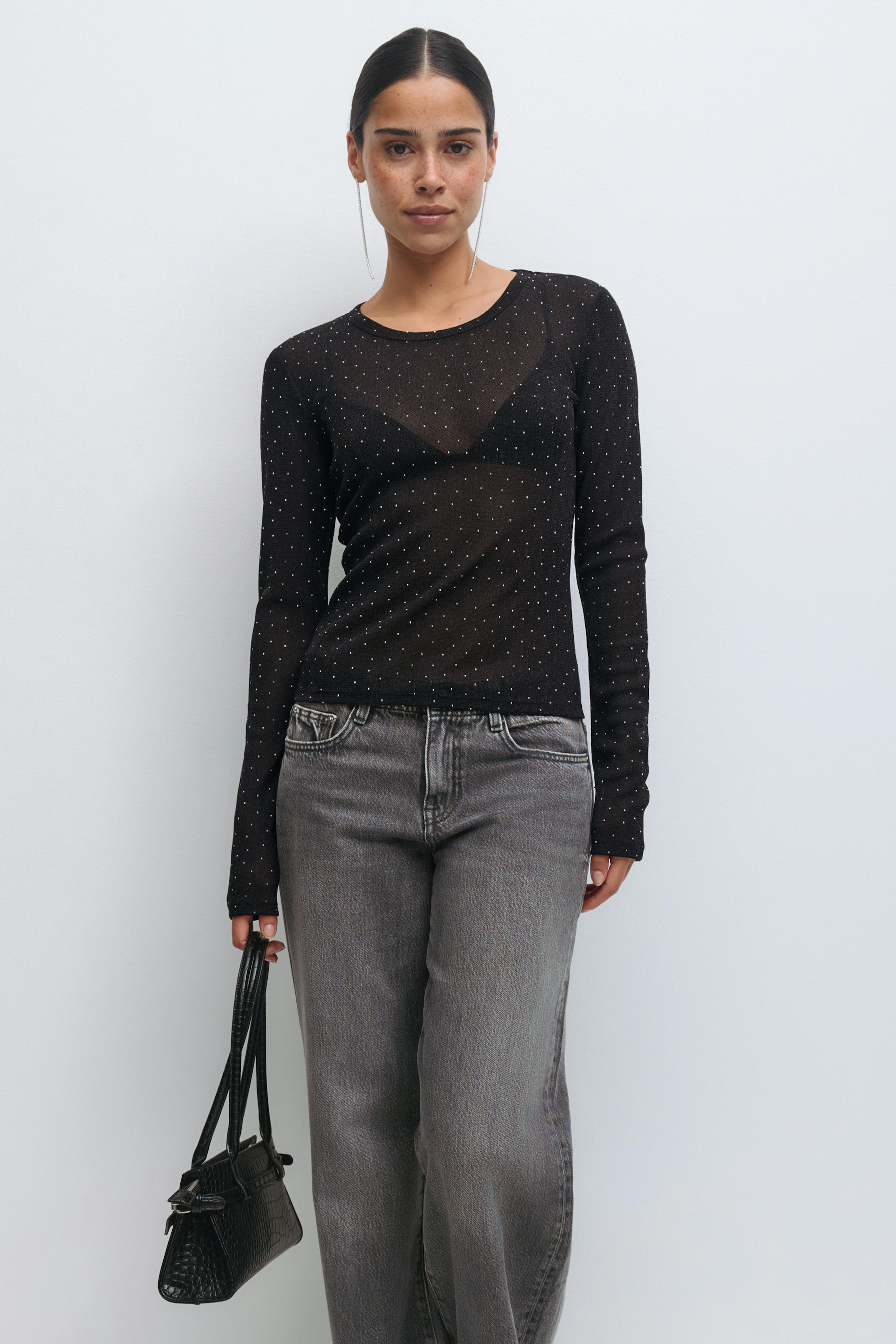Durchsichtiges schwarzes langärmeliges Oberteil mit kleinen Metallic-Punkten über einem schwarzen Triangel-Bralette, verblasste graue High-Waist-Jeans mit geradem Bein, kleine schwarze Handtasche mit Krokoprägung und lange Ohrringe.