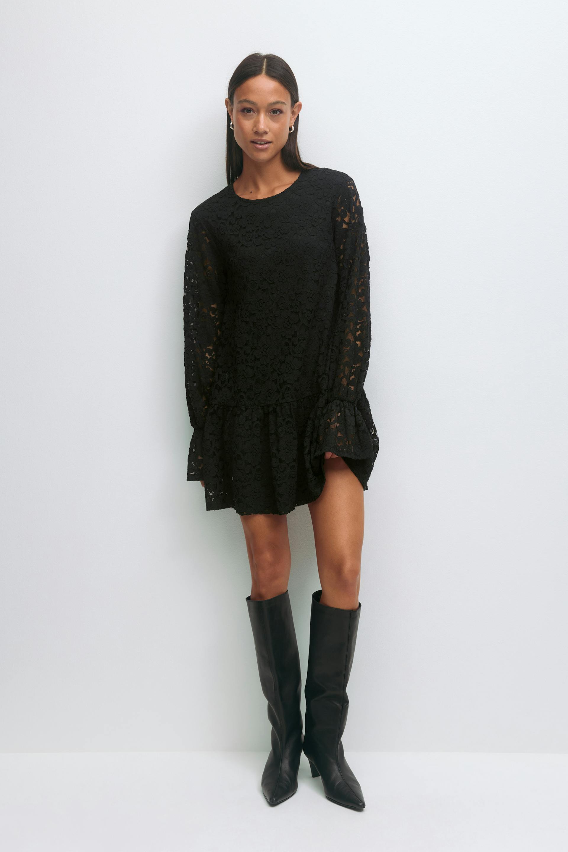 Black floral-lace long-sleeve mini dress with ruffled hem and flared cuffs; black pointed-toe knee-high leather boots.