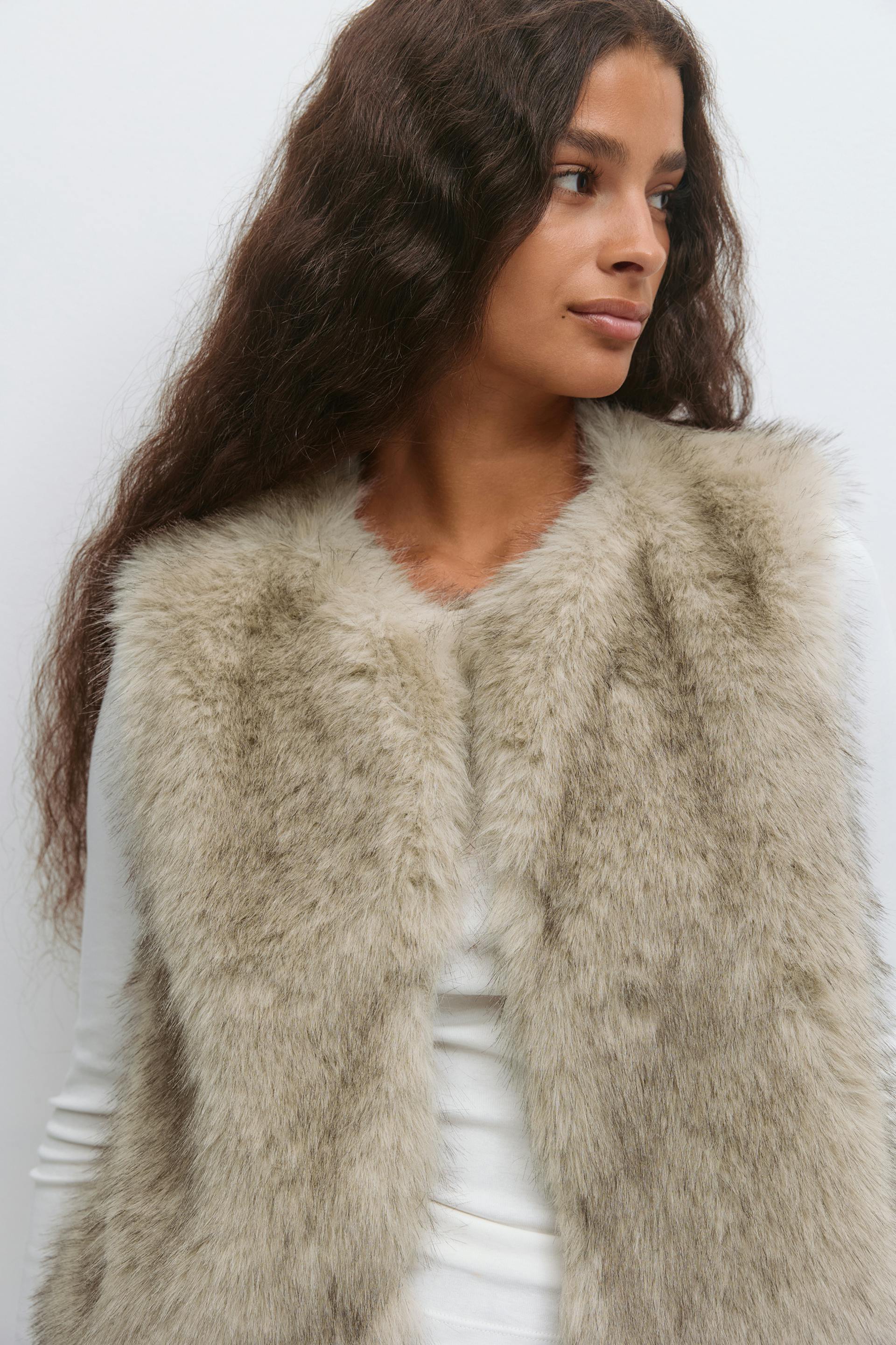 Beige-gray faux-fur sleeveless vest with an open front, layered over a white long-sleeve top.