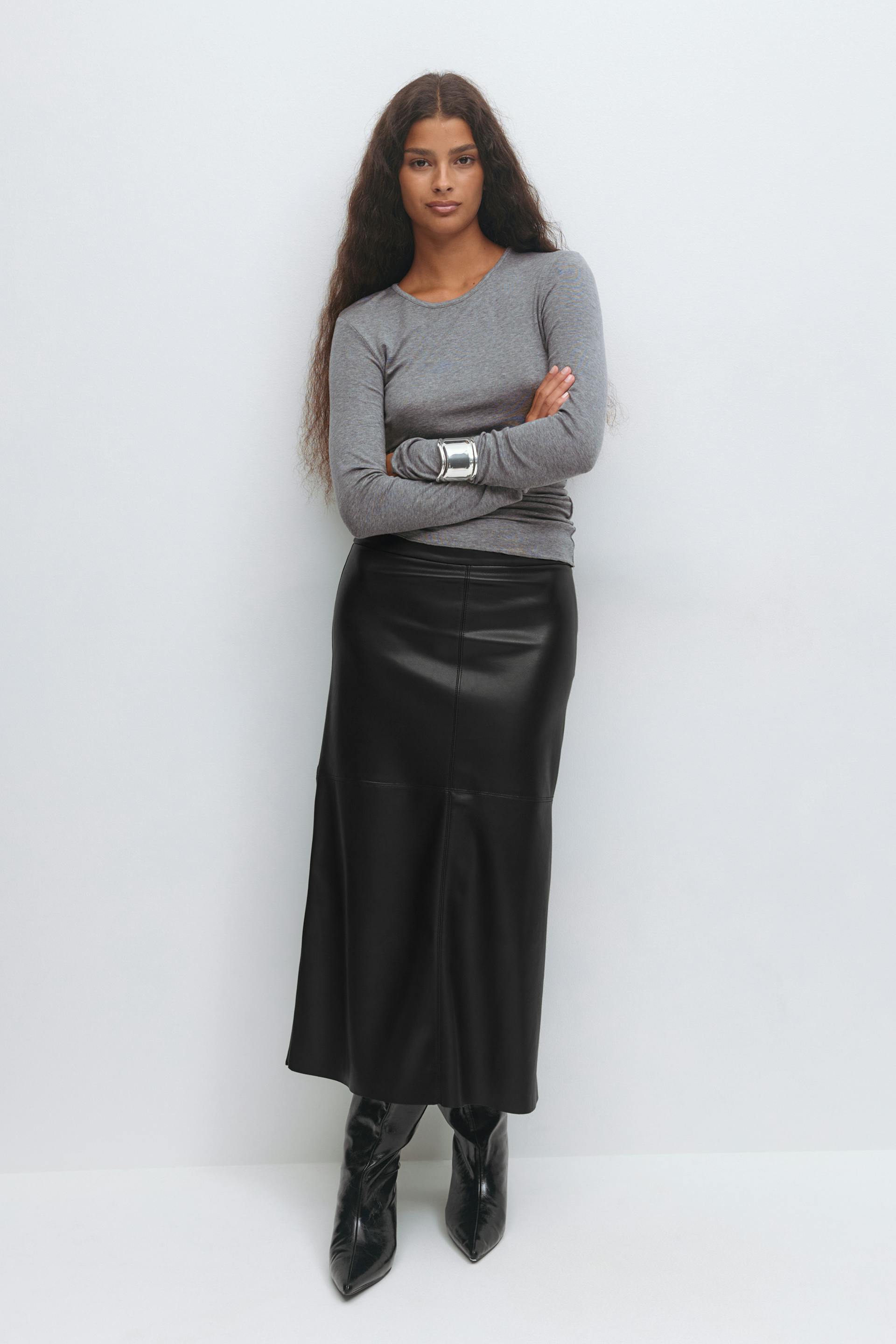 Grey fitted long-sleeve crew-neck top, black leather-look midi skirt with seamed panels, black pointed-toe high-shine boots, and a wide silver cuff bracelet.