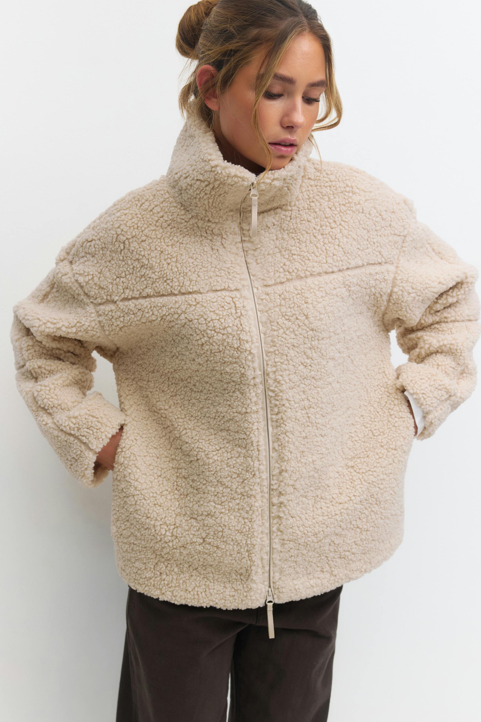 Cremefarbene Sherpa-Zip-up-Jacke mit hohem Trichterkragen, Seitentaschen und langen Ärmeln, kombiniert mit einer dunkelbraunen Hose.