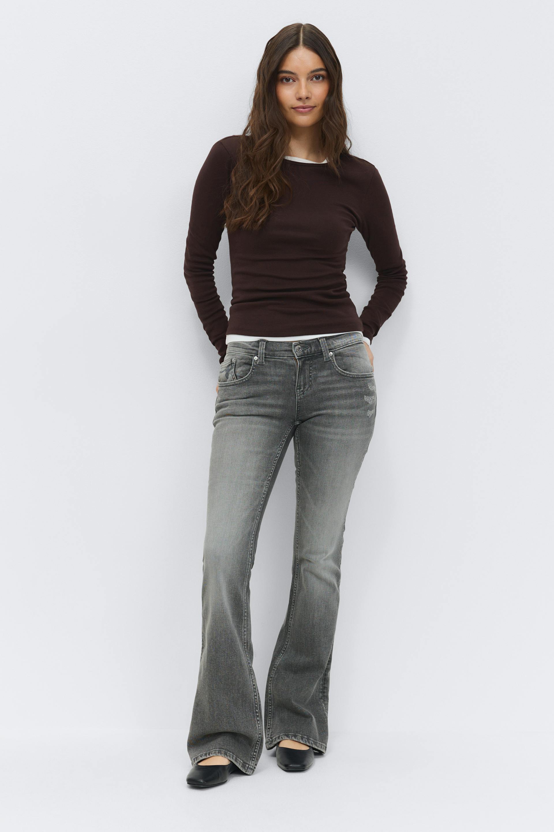 Fitted long-sleeve dark brown crewneck top layered over a white undershirt, grey faded bootcut jeans, and black low-heeled ballet flats.