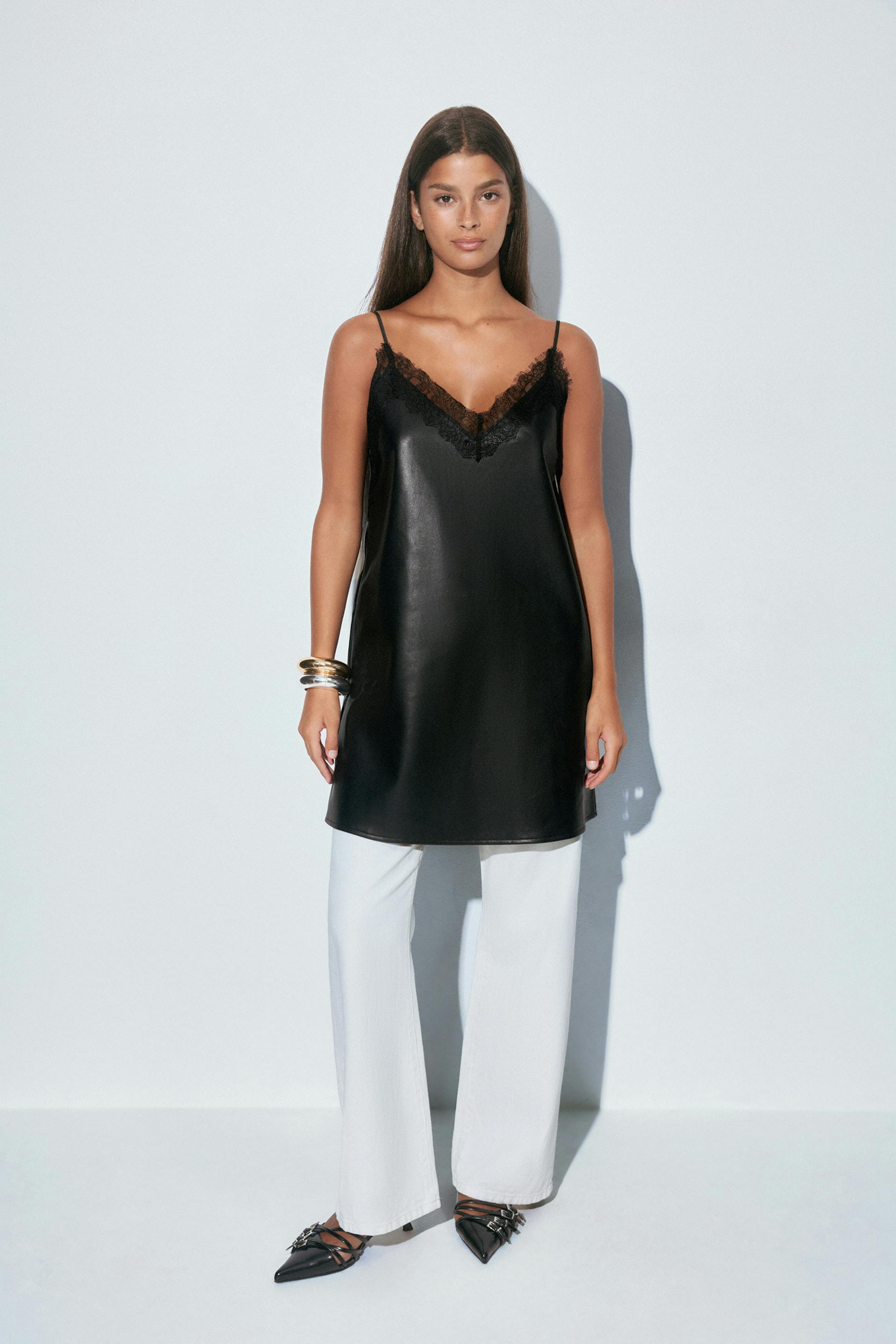 Black faux‑leather camisole slip dress with lace‑trim V‑neck and spaghetti straps layered over white wide‑leg trousers, paired with pointed black strappy flats with metal buckle details and stacked metallic bangles.