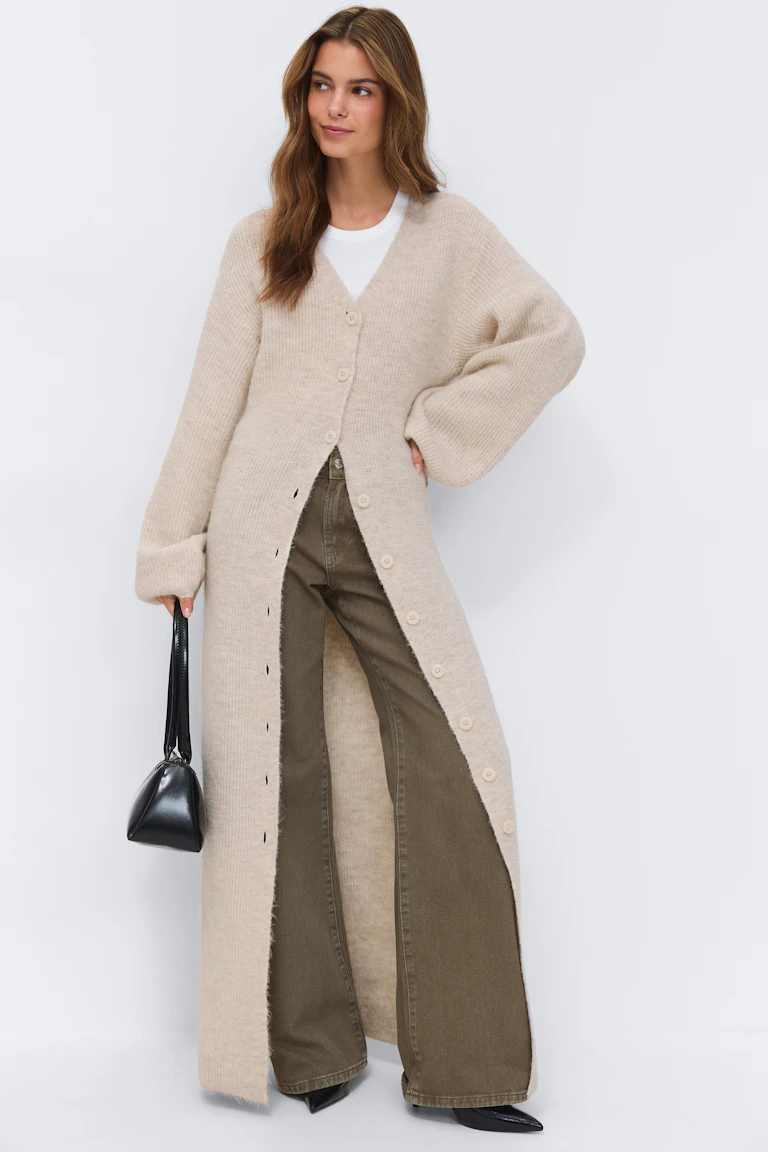 Beigefarbenes Maxikleid aus Rippstrick mit Knopfleiste, V-Ausschnitt und langen Ballonärmeln, kombiniert mit dunkelbraunen, niedrighackigen T-Strap-Pumps.