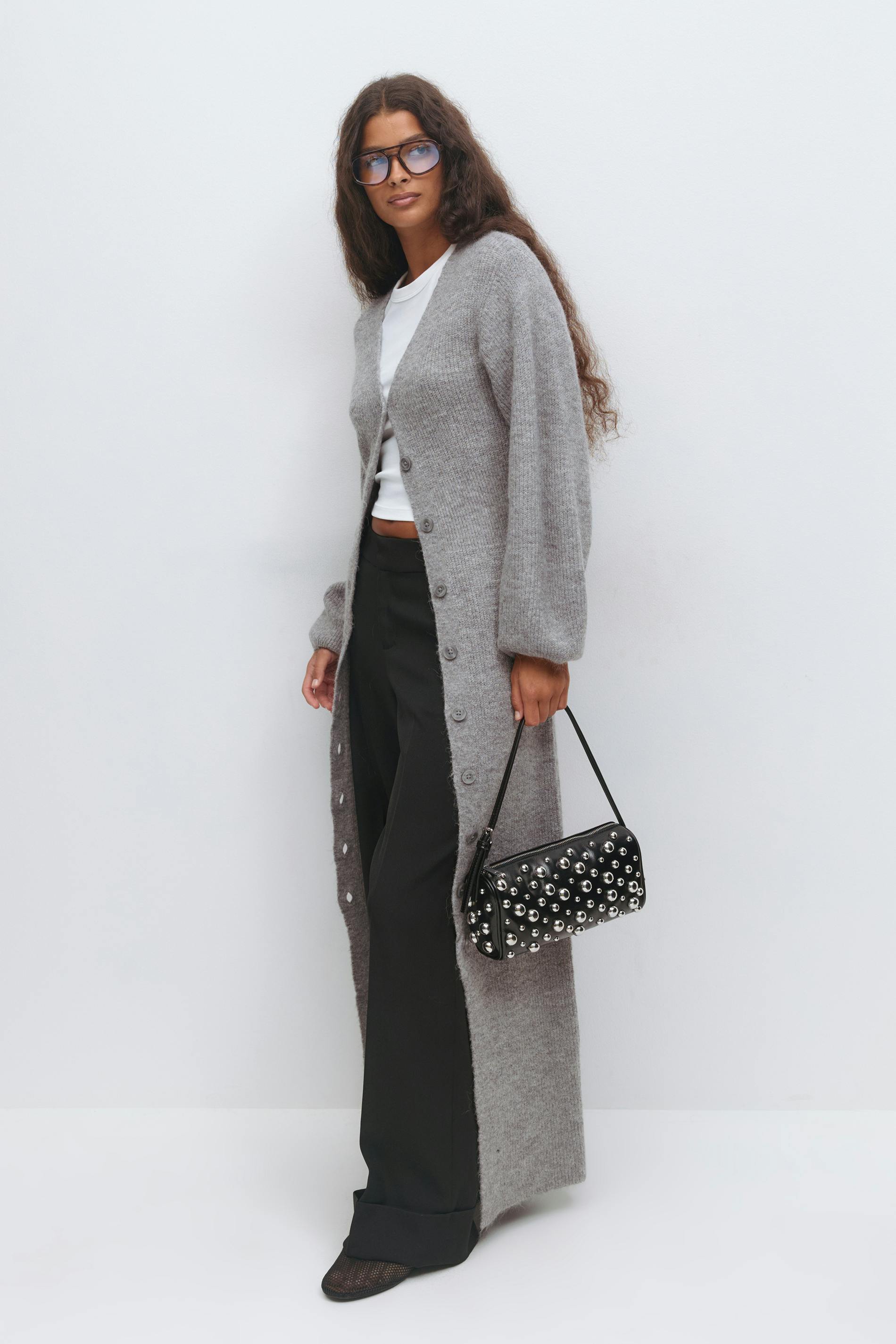 Model wearing a long grey button-front cardigan over a white cropped T‑shirt, black wide-leg trousers, a black studded shoulder bag, black pointed flats and blue-tinted glasses.