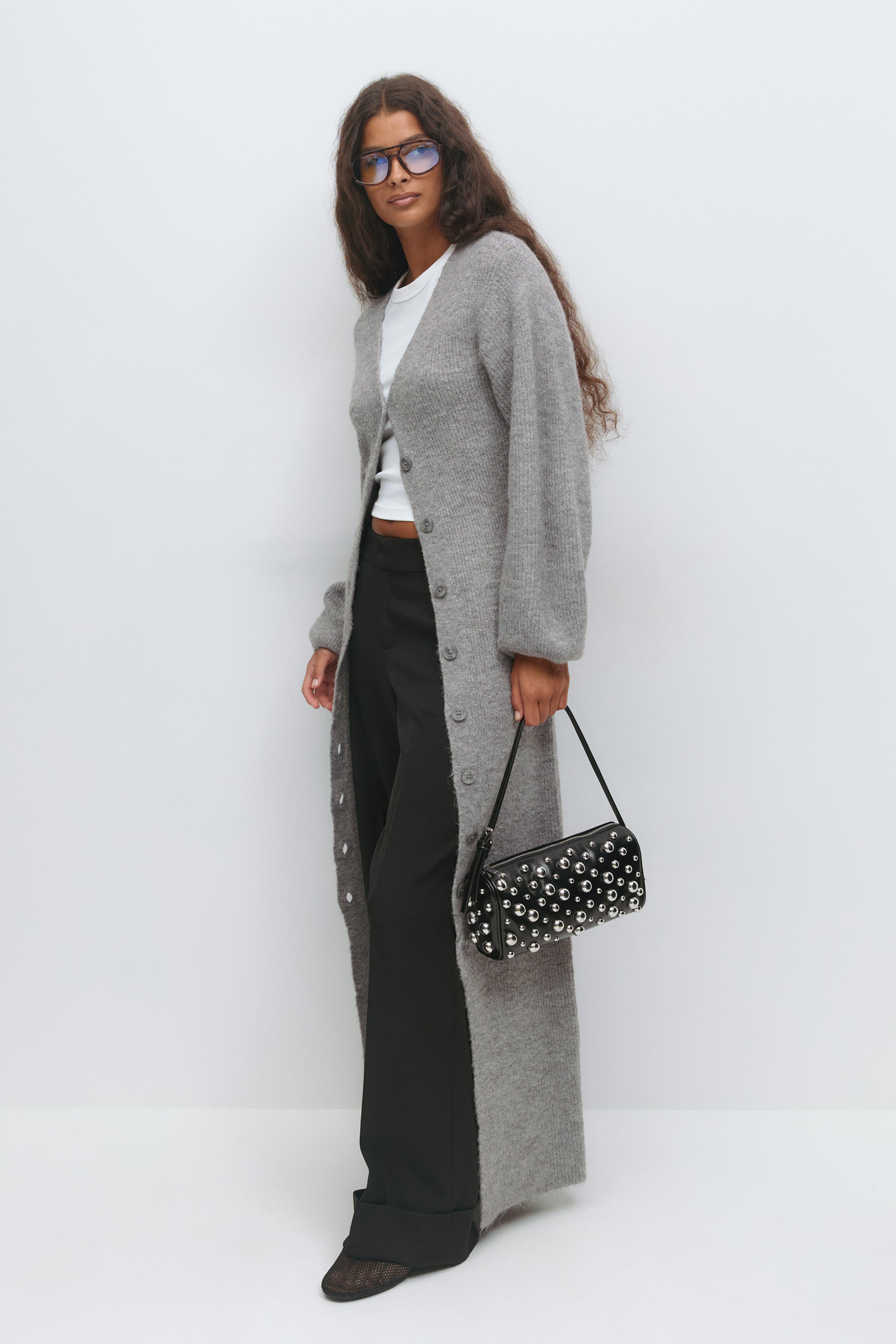 Model wearing a long grey button-front cardigan over a white cropped T‑shirt, black wide-leg trousers, a black studded shoulder bag, black pointed flats and blue-tinted glasses.