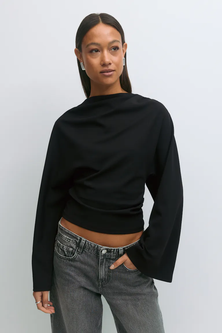 Een zwarte gedrapeerde top met lange mouwen, een boothals en een kortere zoom, gecombineerd met een vervaagde grijze jeans met vijf zakken; afgewerkt met zilveren rechthoekige oorbellen en zilveren ringen.