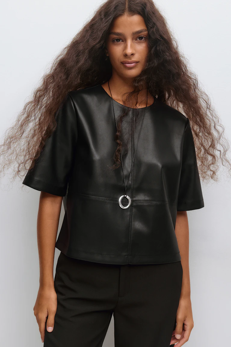 Top boxy noir à manches courtes en similicuir avec coutures horizontales, associé à un pantalon de tailleur noir et à un long collier à cordon noir avec pendentif circulaire argenté.