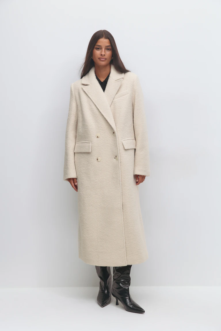Double-breasted cream wool long coat with notch lapels and flap pockets, worn over a black top; paired with knee-high black patent leather pointed-toe heeled boots.