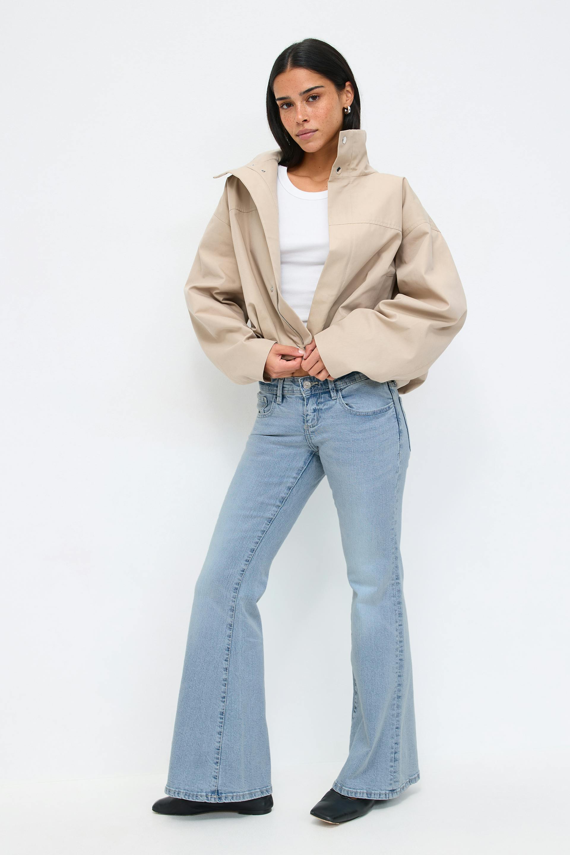 Beige oversized high-collar jacket with snap closures over a white crew-neck tank, light-wash flared jeans and black pointed-toe ankle shoes.