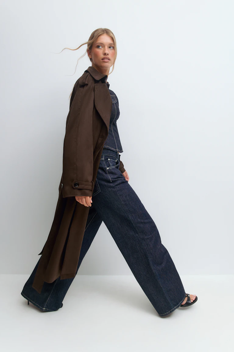 Long trench-coat marron foncé drapé sur les épaules, top ajusté à corset en denim indigo foncé, jean large taille haute indigo foncé avec surpiqûres contrastées, et sandales à talons noires à lanières.