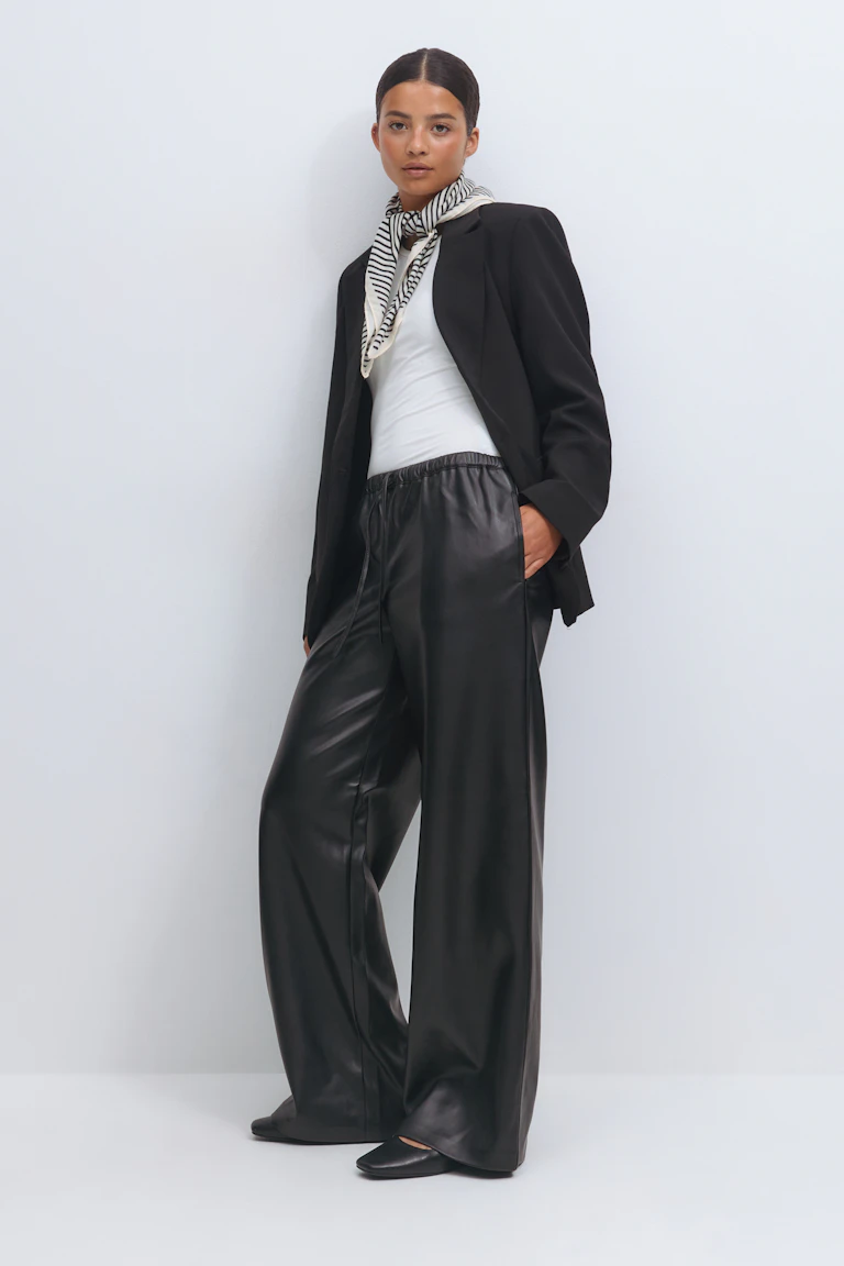 Black tailored blazer worn over a white fitted top, a black-and-white striped neck scarf, black wide-leg leather-look trousers with elastic waist, and black pointed flats.