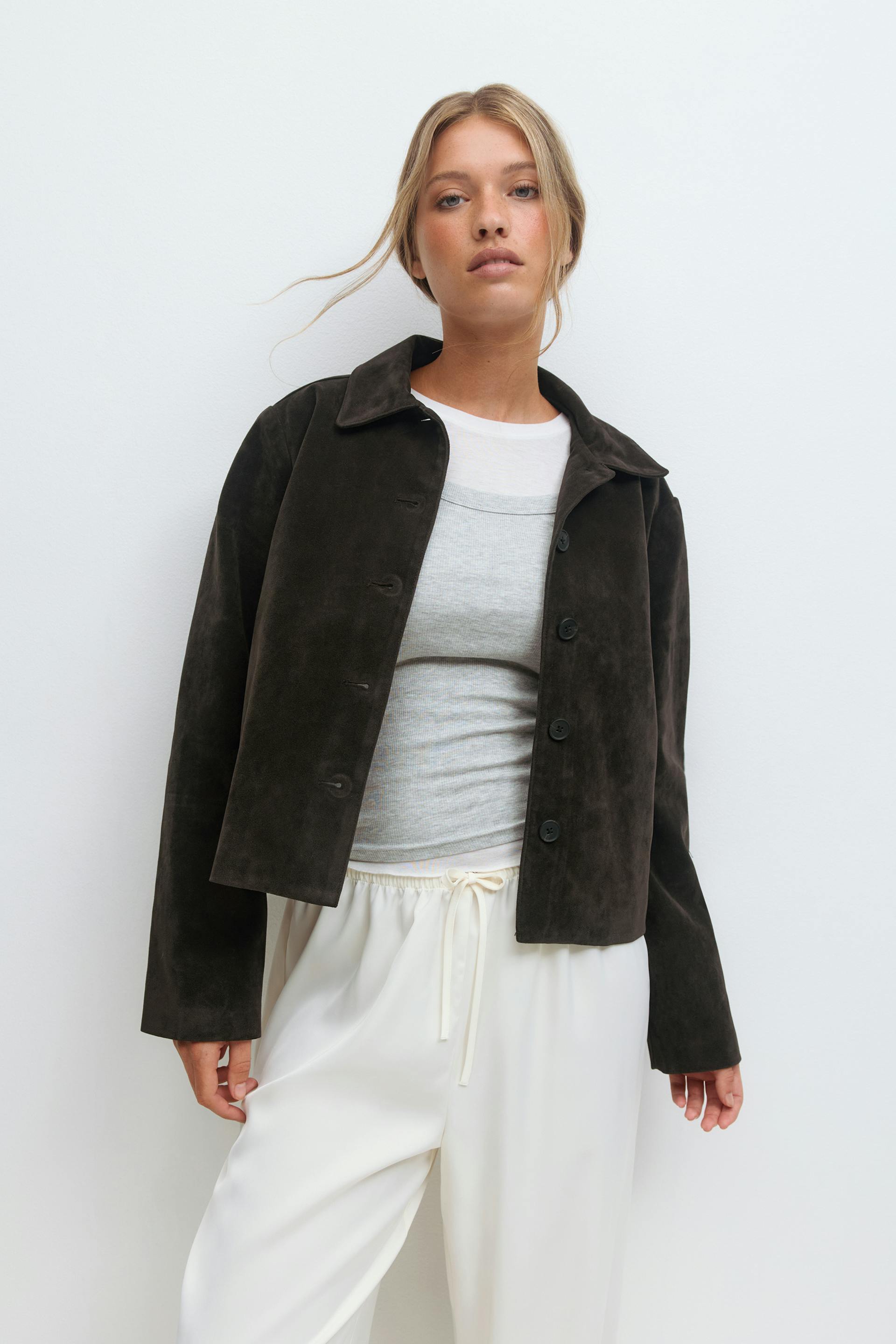 Dark brown suede button-front collared jacket layered over a light gray ribbed tank and white crew-neck tee, paired with ivory drawstring wide-leg trousers.