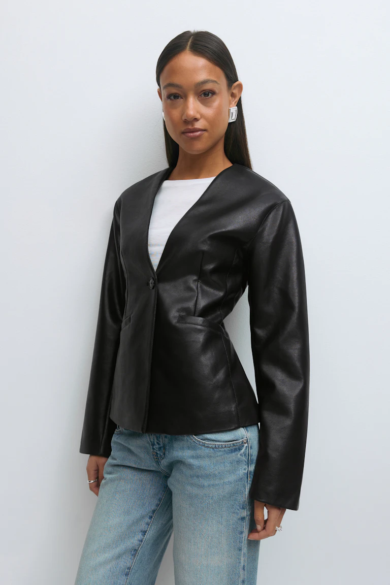 Maßgeschneiderter schwarzer Lederblazer mit einem einzigen Knopf über einem weißen T-Shirt mit Rundhalsausschnitt, kombiniert mit einer hellen Jeans mit geradem Bein; dazu silberne geometrische Ohrringe und Ringe.