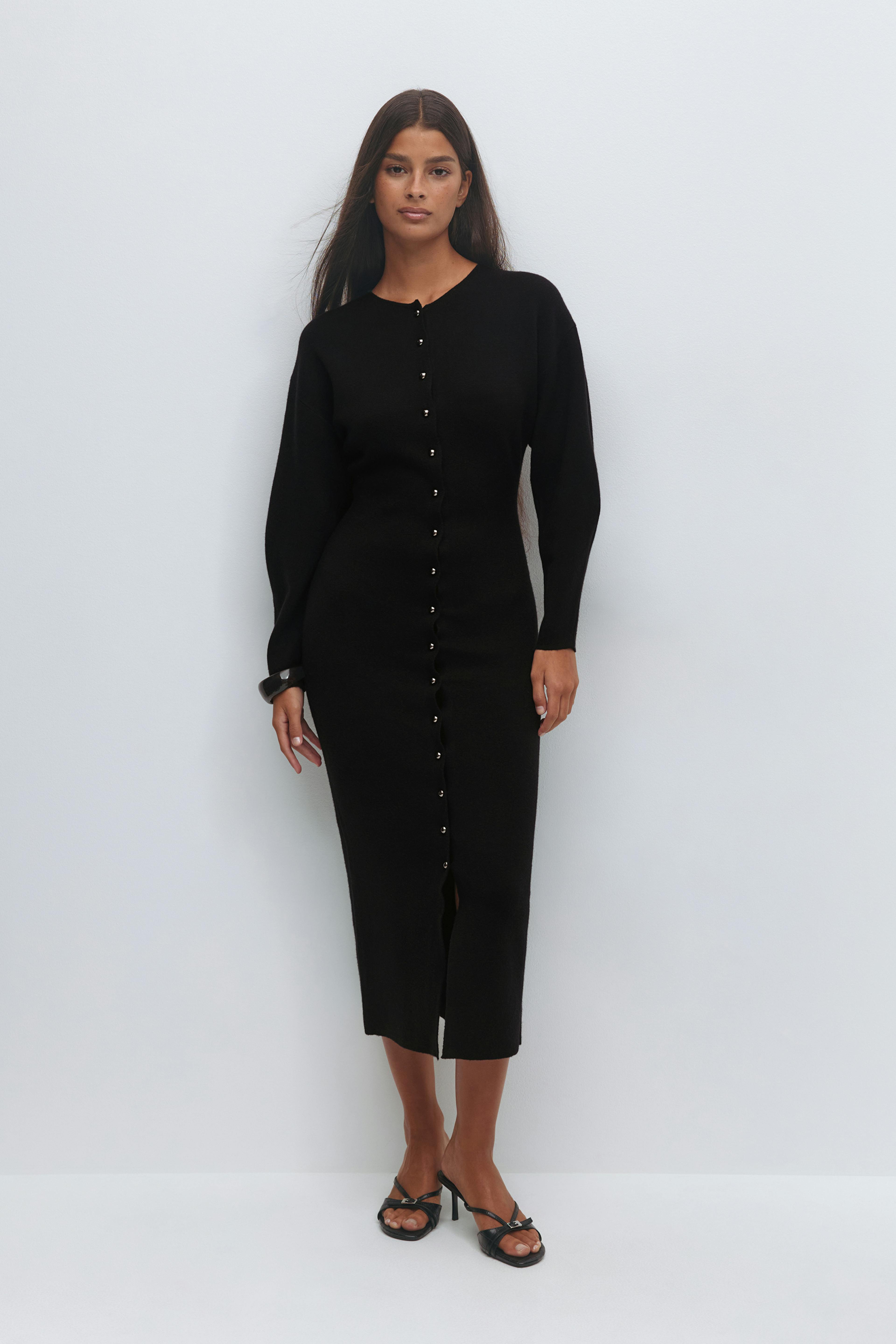 Black long-sleeve button-front midi knit dress with a center front slit, styled with black strappy heeled sandals and a black cuff bracelet.