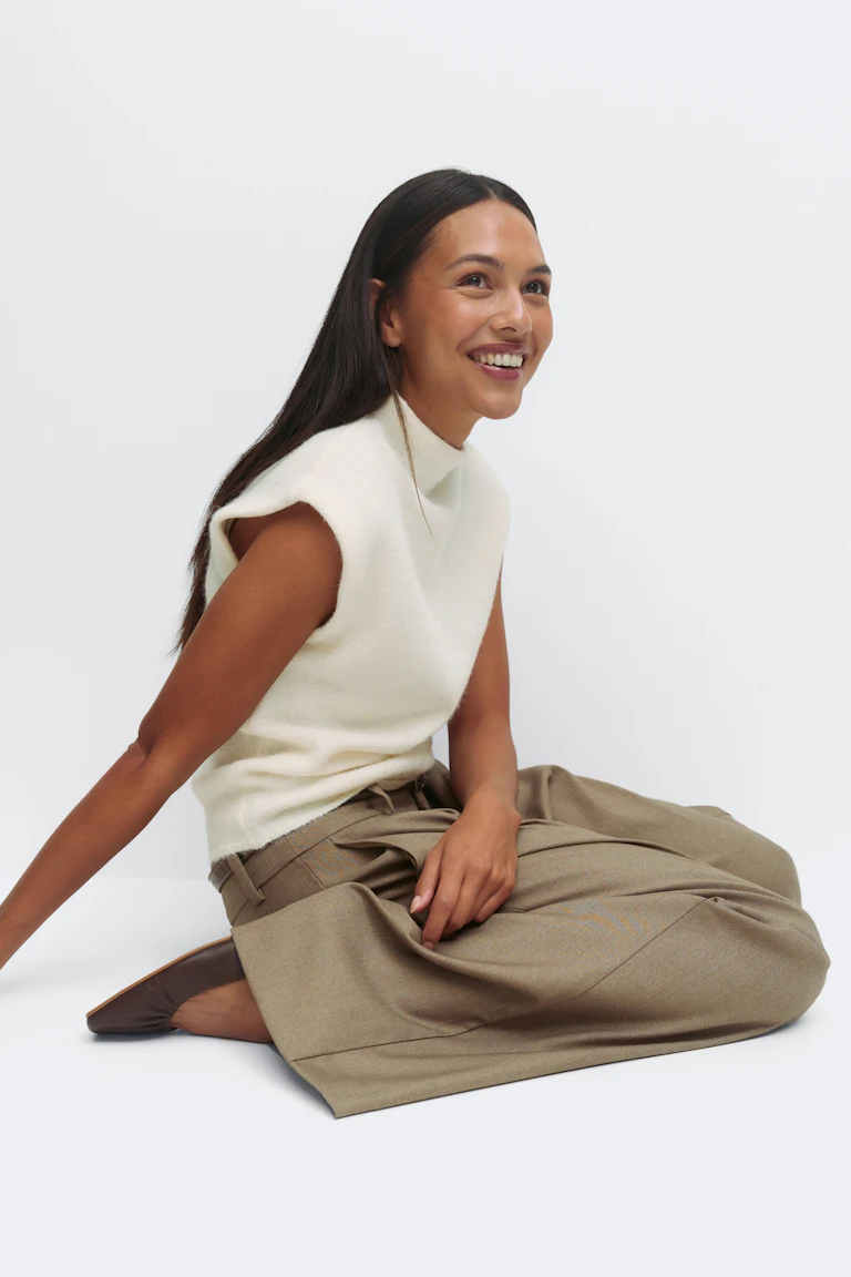 Mouwloze gebreide top met crèmekleurige mock-hals in combinatie met een taupe getailleerde broek met hoge taille en wijde pijpen en donkerbruine puntige leren flats.