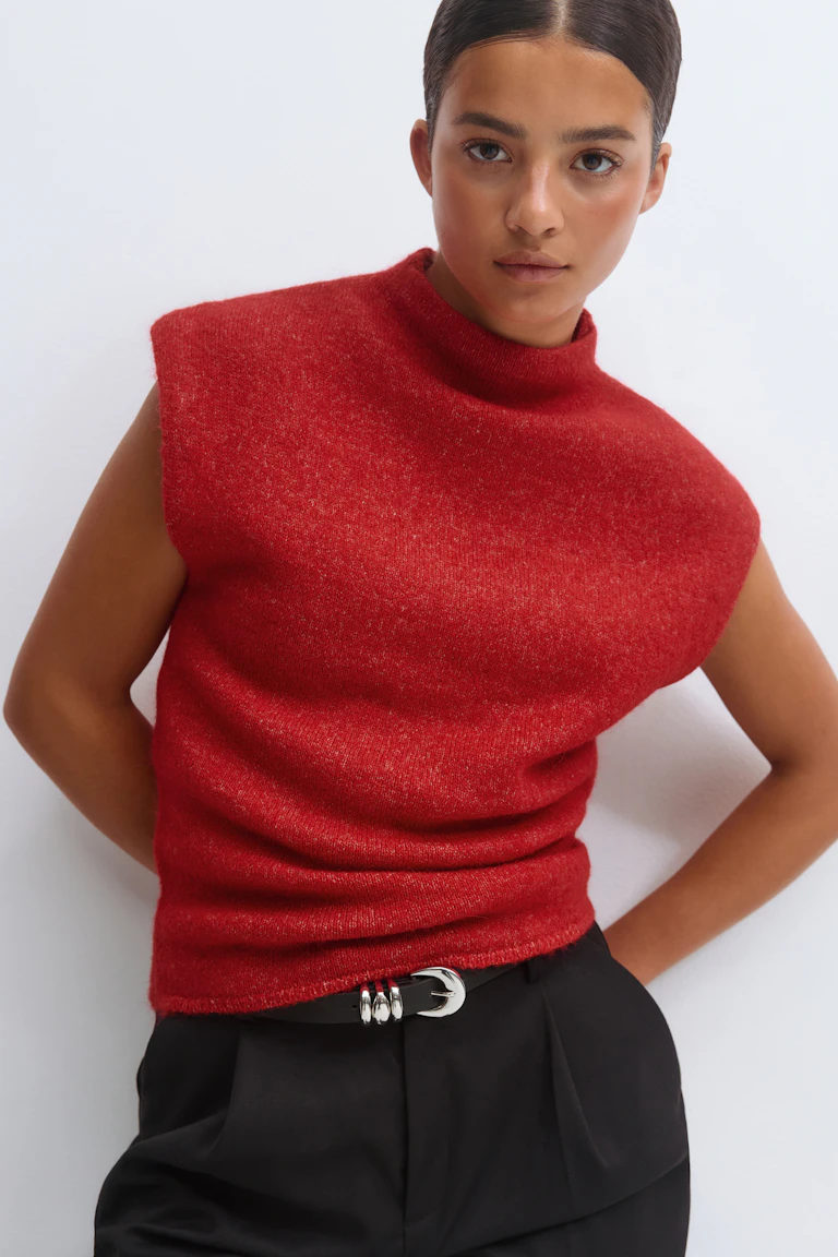 Top en tricot rouge sans manches à col maillé, associé à un pantalon de tailleur noir et à une ceinture noire à boucle en argent poli.