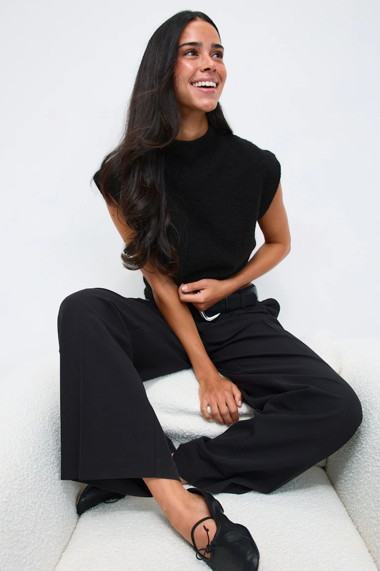 Sort strikket top uden ærmer med mock-neck, højtaljede sorte bukser med vide ben og sort bælte med sølvspænde og sorte ballerinaer i læder med bindebånd.