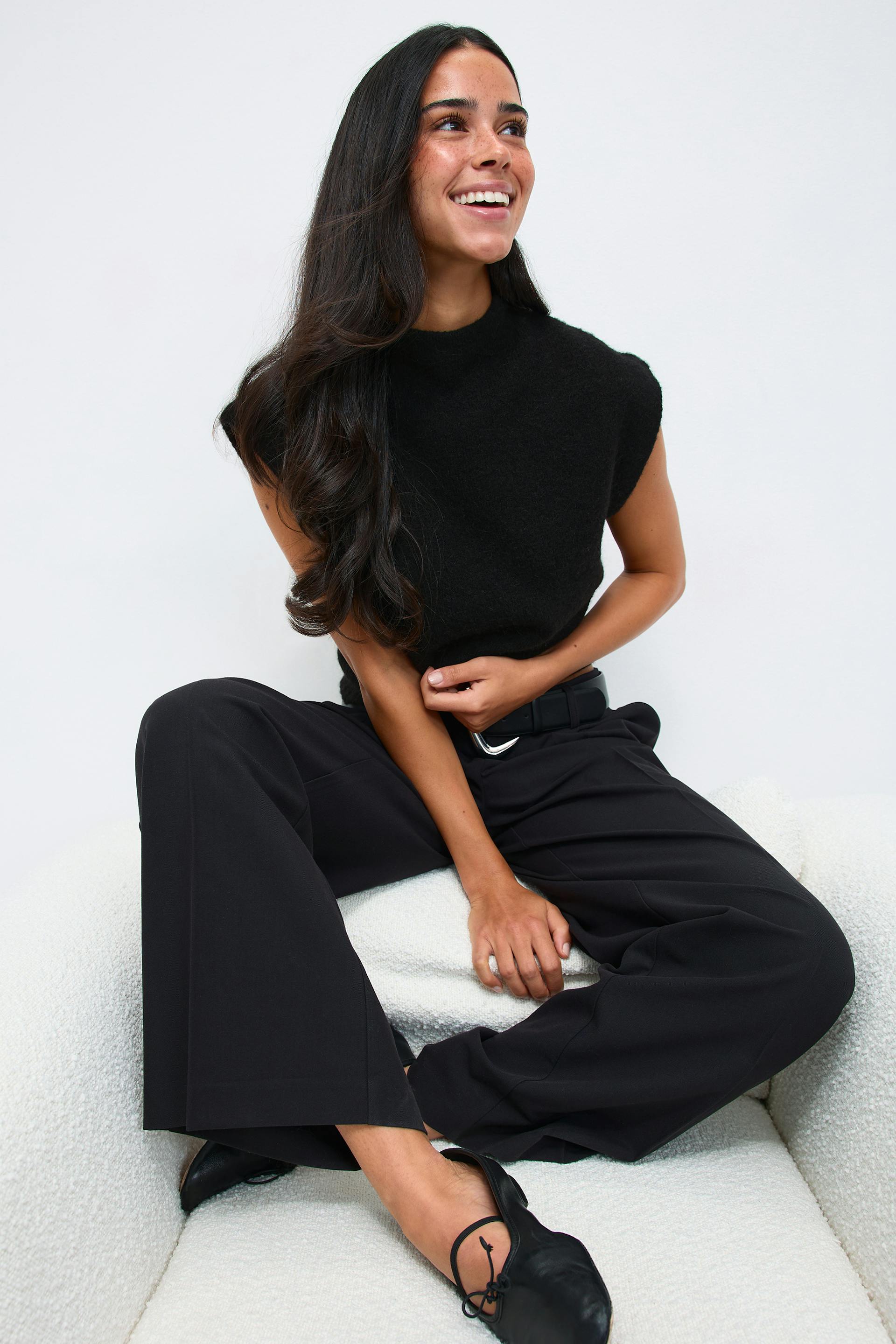 Black sleeveless mock‑neck knit top, high‑waisted black wide‑leg trousers with a black belt and silver buckle, and black leather ballet flats with tie detail.