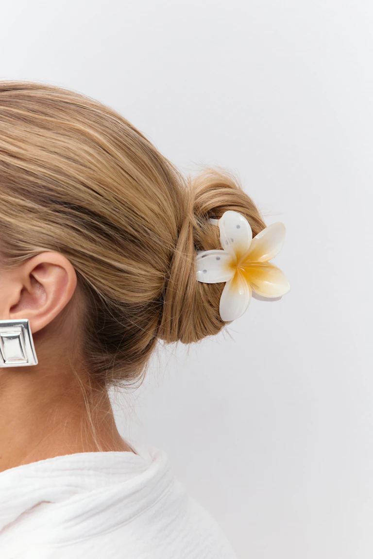 Hvit og gul plumeria-hårspenne som fester en lav knute, overdimensjonerte firkantede sølvøreringer og hvit bluse med struktur.