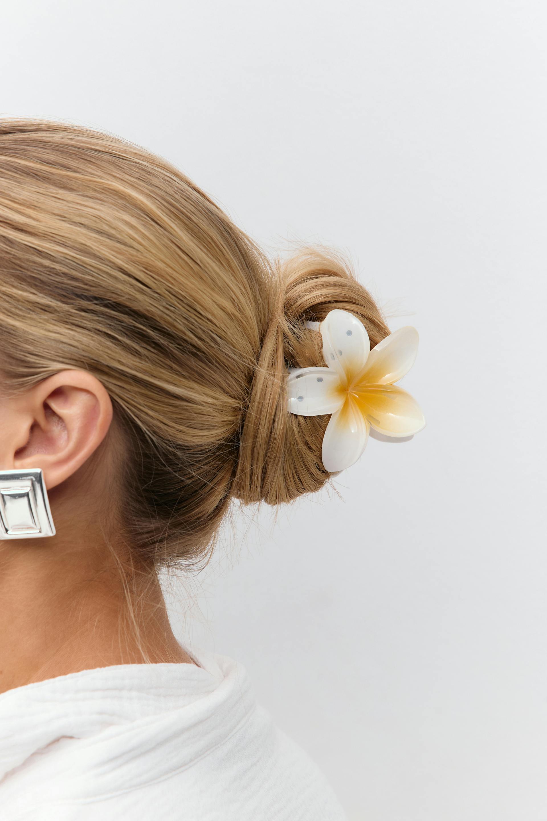 White-and-yellow plumeria hair clip securing a low bun, oversized square silver earring, and white textured blouse.