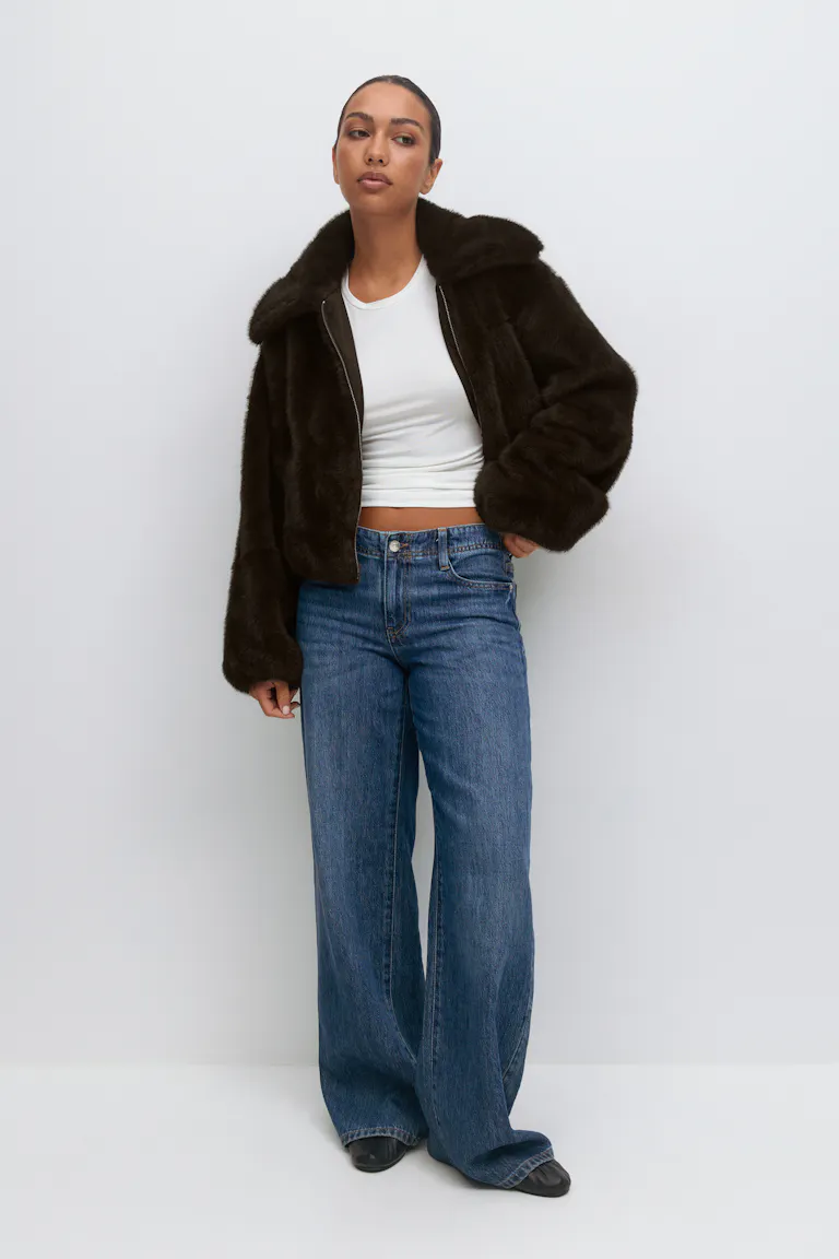 Dunkelbraune Kunstpelzjacke mit Reißverschluss und großem Kragen, weißes T-Shirt mit Rundhalsausschnitt, blaue Jeans mit hoher Taille und schwarzen Schuhen mit spitzen Zehen.