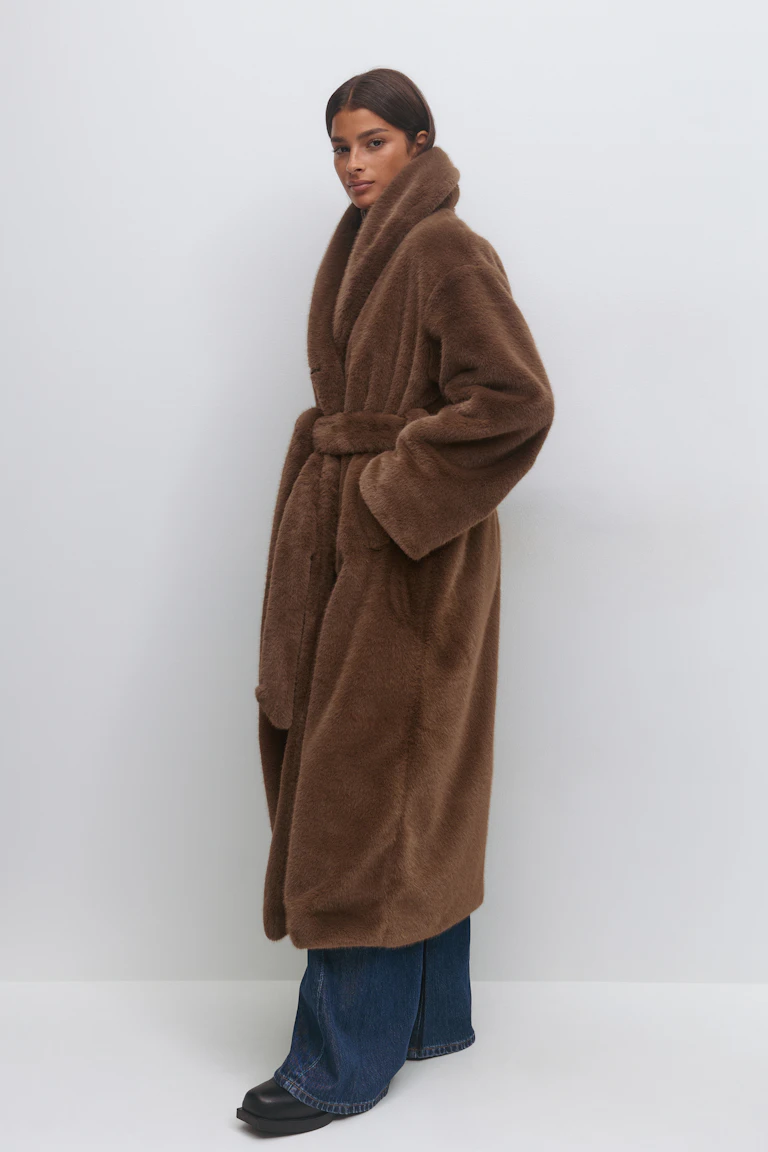 Brown long belted faux-fur coat with shawl collar and long sleeves, paired with dark blue wide-leg jeans and black square-toe boots.