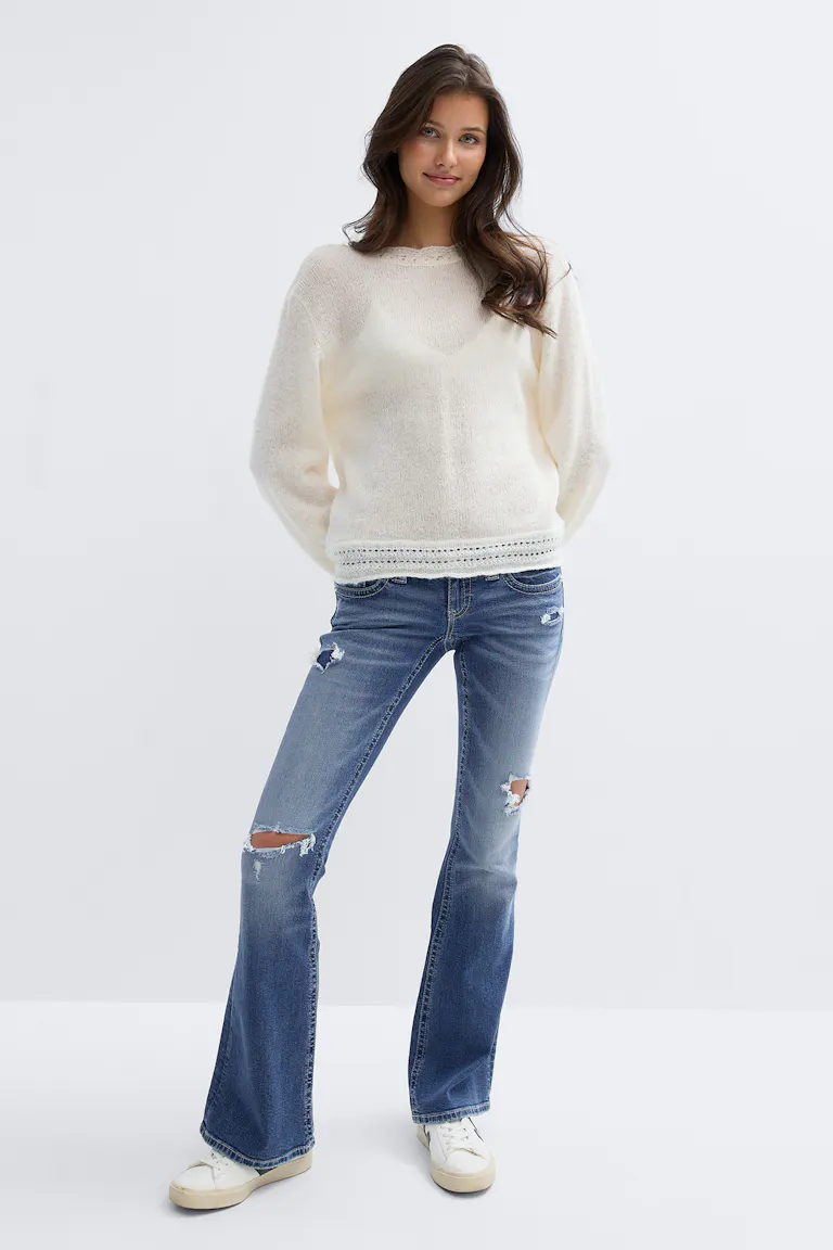 Elfenbensfärgad långärmad lättstickad crewneck-tröja med genombruten fåll, mediumtvättade slitna utsvängda jeans med knäveck och vita sneakers med låg topp.