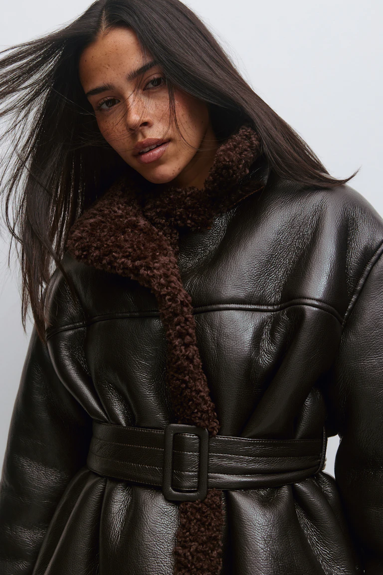 Belted dark brown leather-look coat with curly shearling collar and trim and a rectangular buckle.