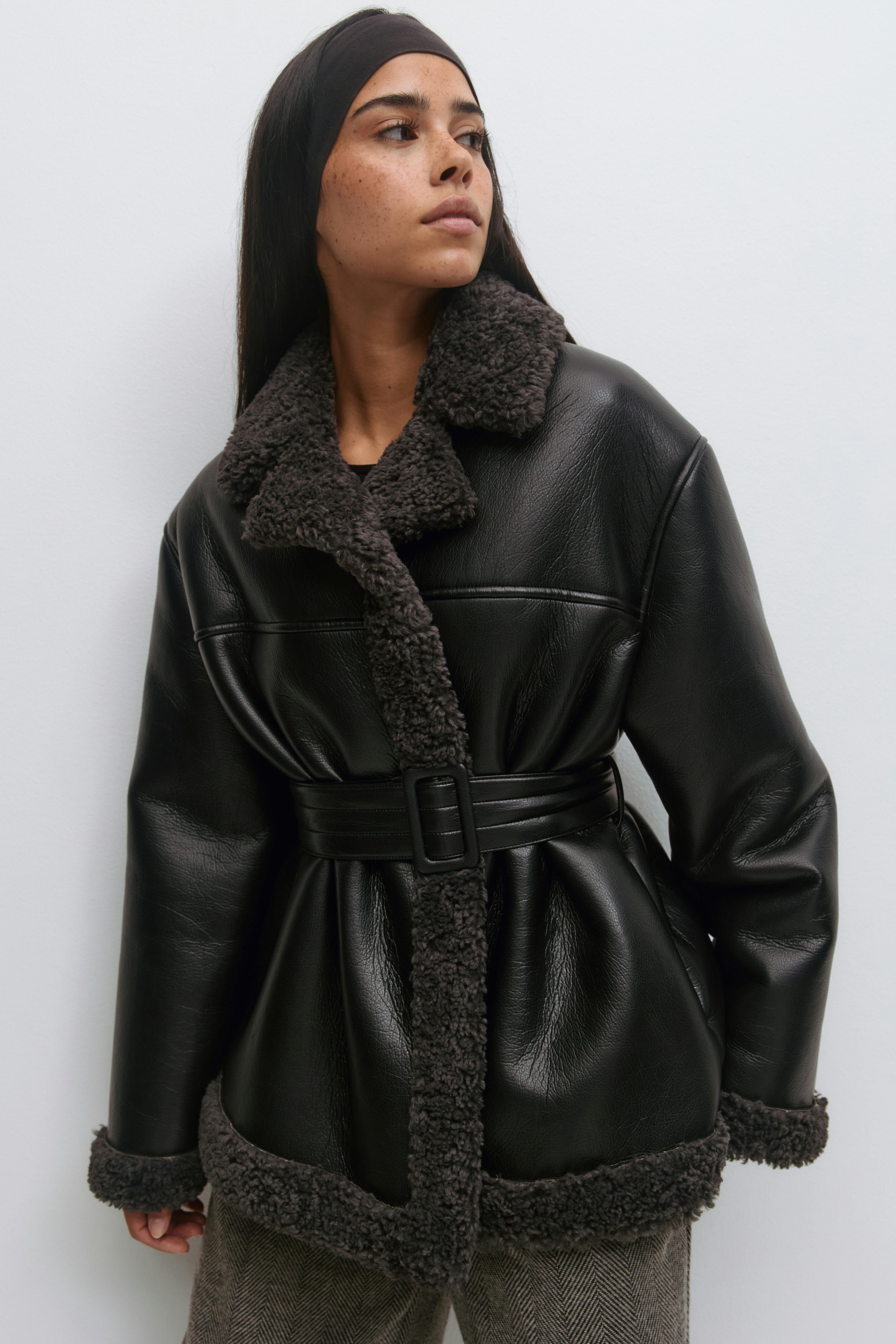 Sort kunstlæderjakke med bælte og trækulsfarvet kunstshearling på krave, stolpelukning, manchetter og forneden, sammen med grå sildebensbukser.