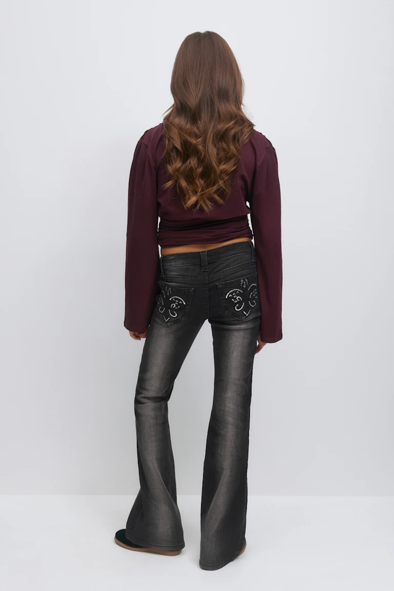 Wearing a long-sleeve burgundy cropped top paired with faded black flared jeans featuring decorative embroidered and studded heart motifs on the back pockets, and black slip-on shoes.