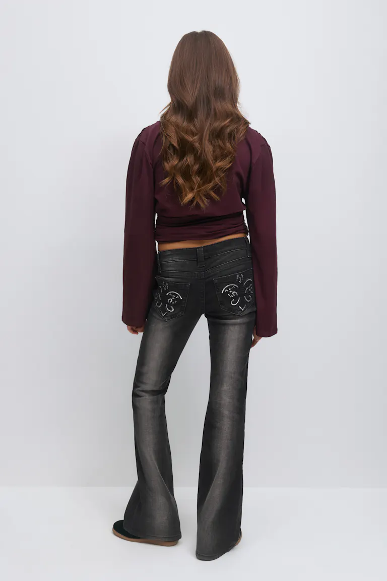 Wearing a long-sleeve burgundy cropped top paired with faded black flared jeans featuring decorative embroidered and studded heart motifs on the back pockets, and black slip-on shoes.