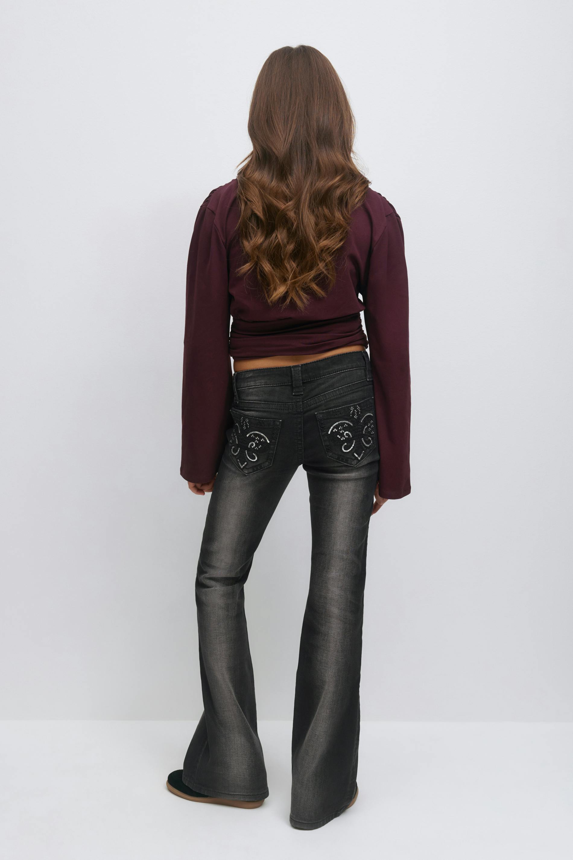 Yllään pitkähihainen viininpunainen cropped-toppi yhdistettynä haalistuneisiin mustiin leveneviin farkkuihin, joiden takataskussa on koristeellisia kirjailtuja ja nastoitettuja sydänaiheita, ja mustat slip-on-kengät.