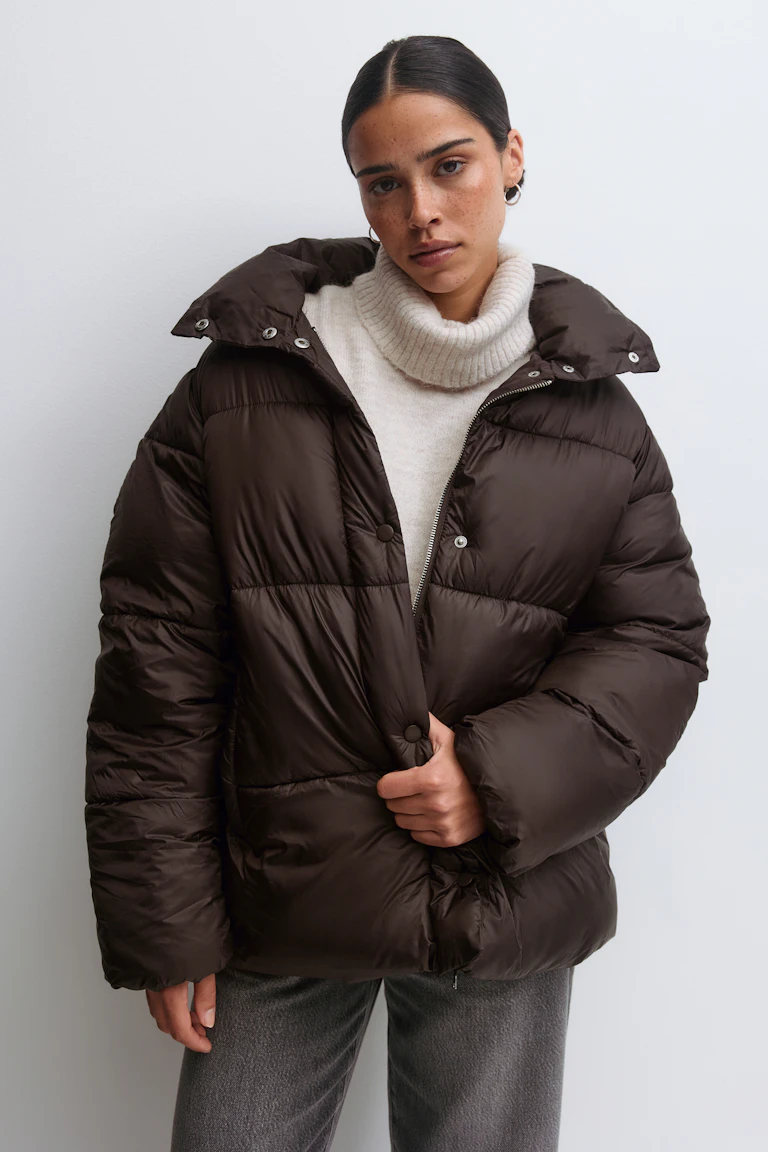 Donkerbruin oversized gewatteerd pufferjack met hoge kraag en drukknoop-/ritssluiting, crèmekleurige geribde coltrui en grijze broek met rechte pijpen.
