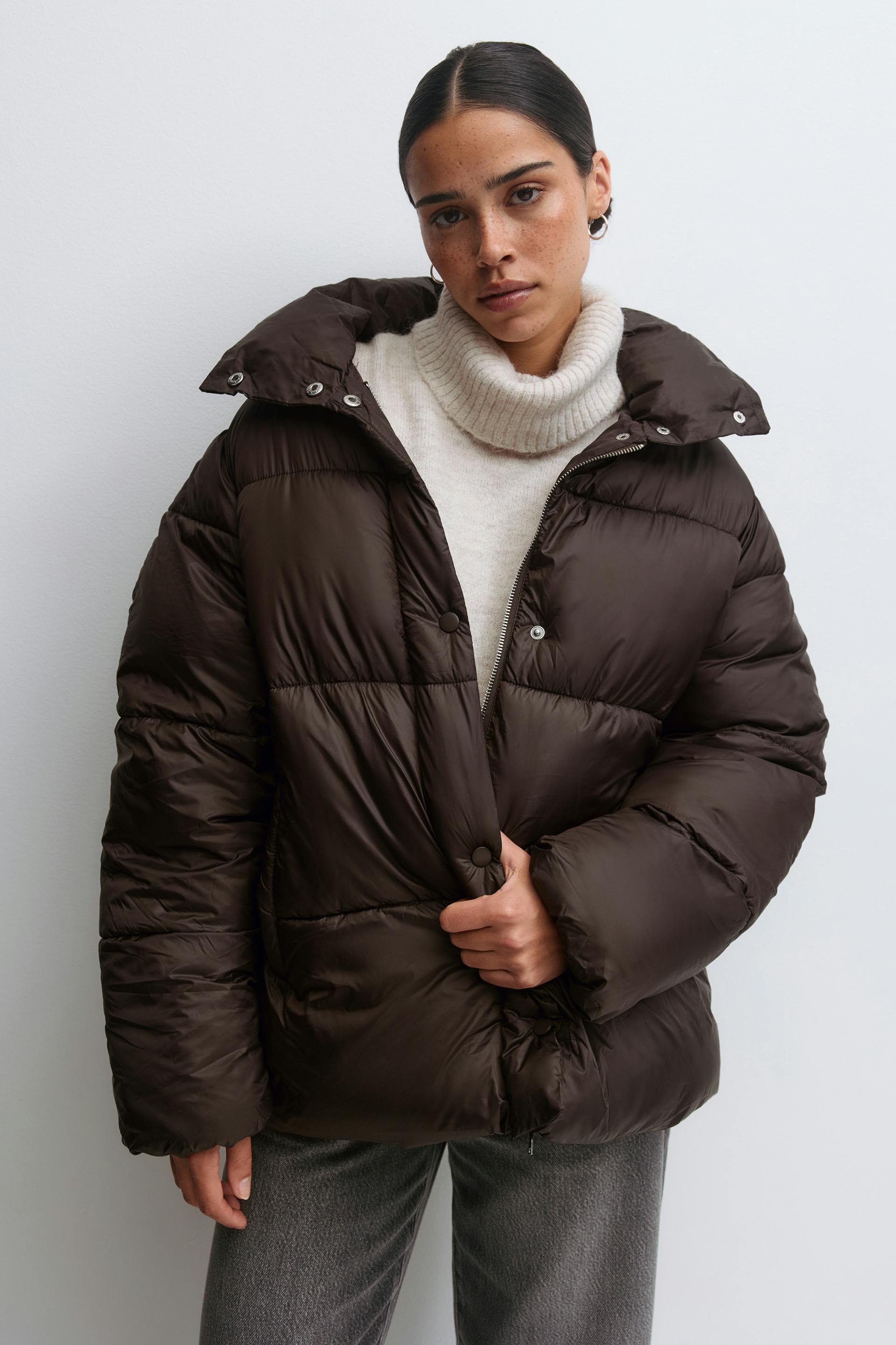Dark brown oversized quilted puffer jacket with high collar and snap/zip closure, cream ribbed turtleneck sweater, and grey straight-leg trousers.