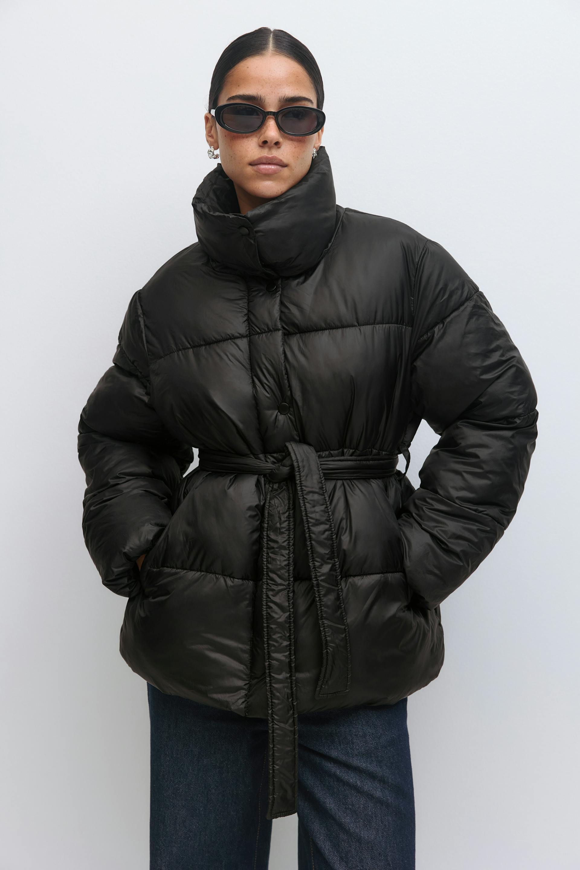 Black belted puffer jacket with high funnel collar and horizontal quilting, styled with dark blue straight-leg jeans, black oval sunglasses, and small silver earrings.