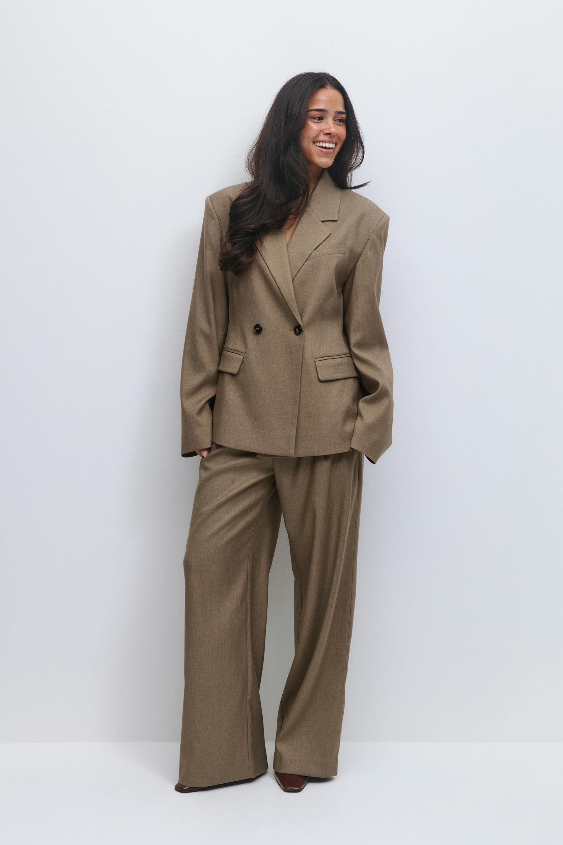 Zweireihiger Blazer in Taupe mit eingekerbtem Revers, Pattentaschen und zwei dunklen Knöpfen, kombiniert mit einer passenden Hose mit weitem Bein und braunen Schuhen mit spitzen Zehen.