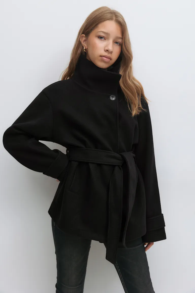 Black belted wool coat with high stand collar, single-button front and cuffed long sleeves, paired with dark washed slim jeans and small gold hoop earrings.