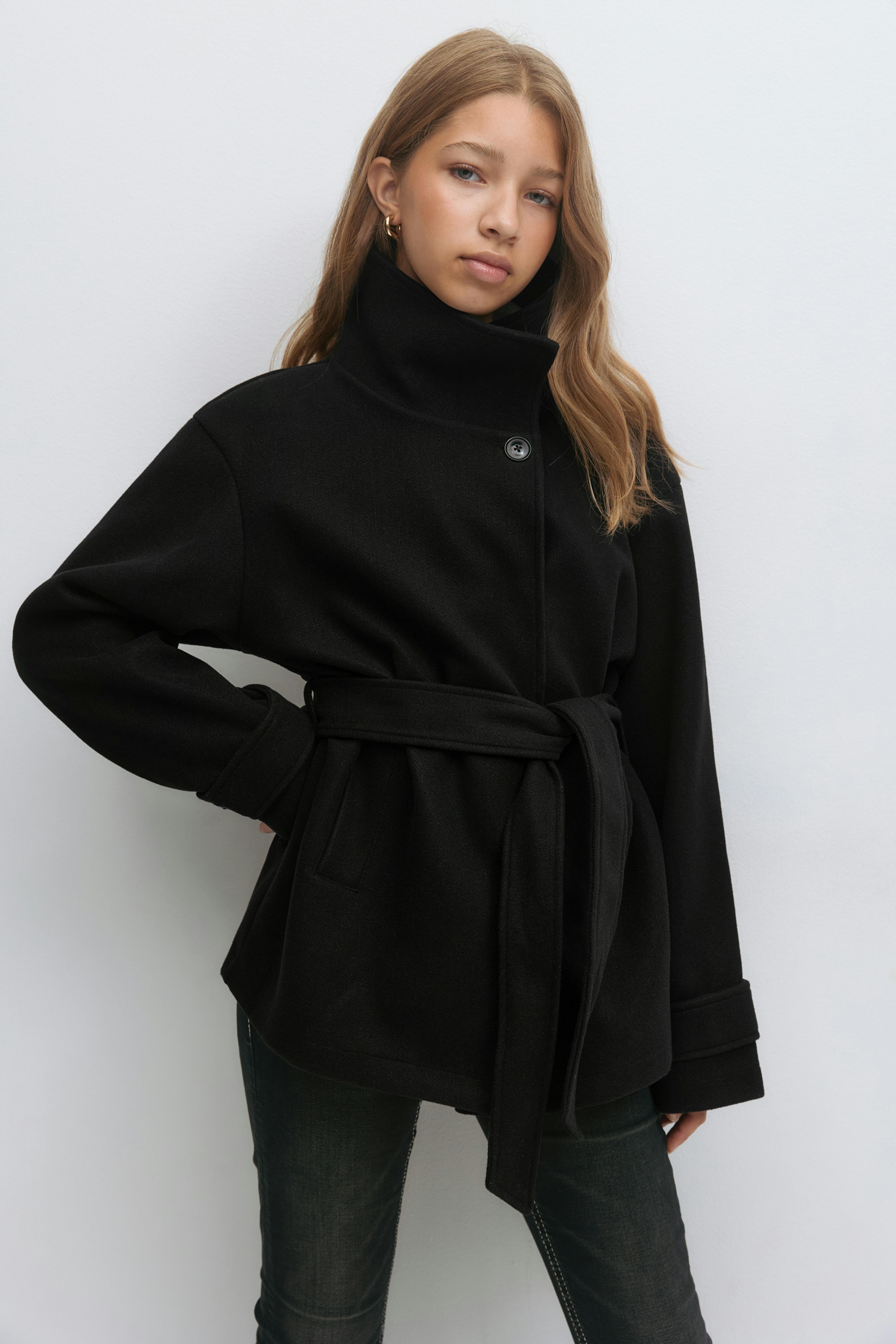 Manteau en laine noir ceinturé avec col montant, simple boutonnage et manches longues à revers, associé à un jean slim délavé et à de petites boucles d'oreilles dorées.