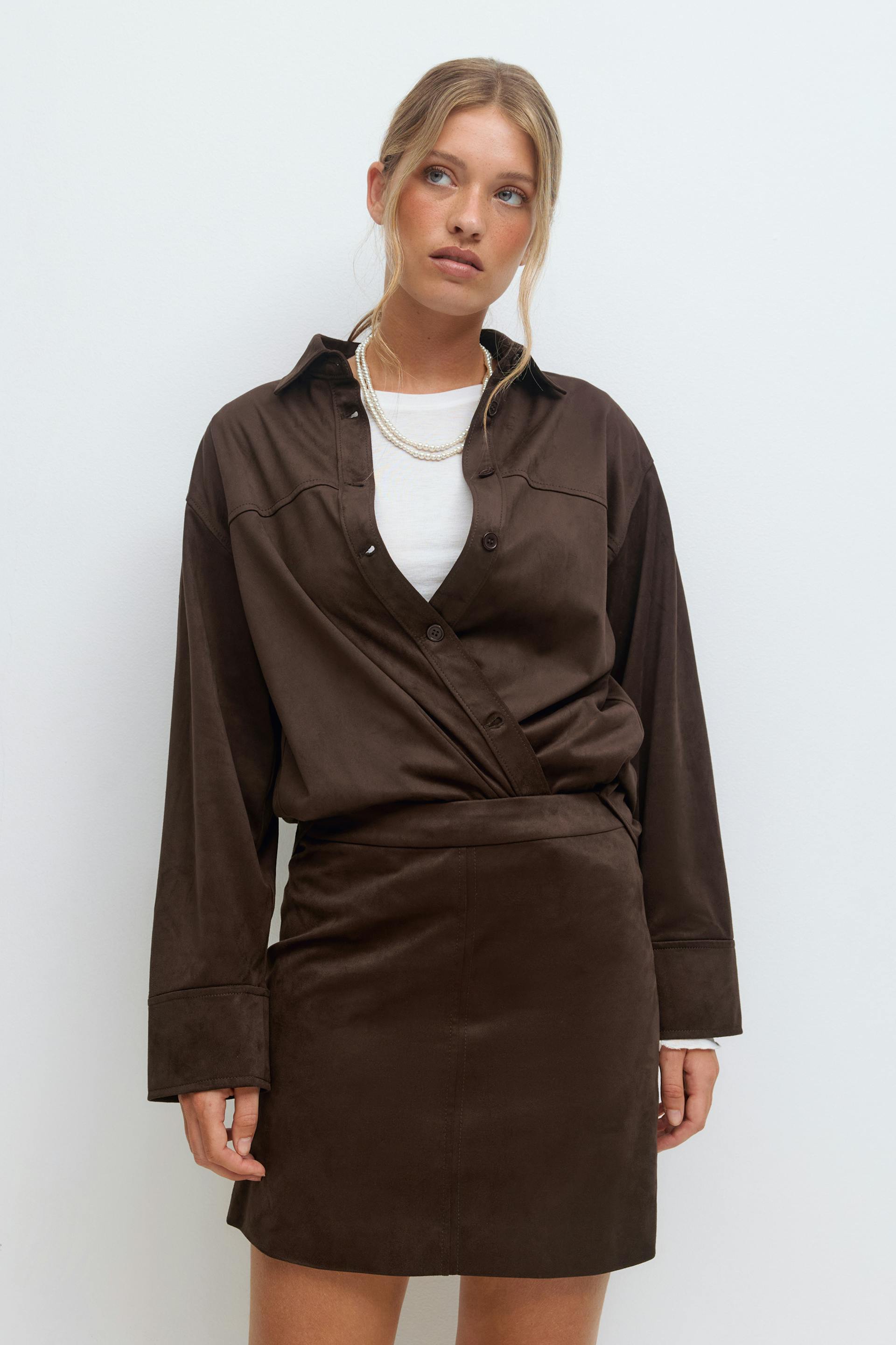 Mørkebrun langermet skjorte i slå-om-stil med store mansjetter over en hvit t-skjorte med rund hals, sammen med et matchende mørkebrunt miniskjørt og lag på lag med perlekjeder.