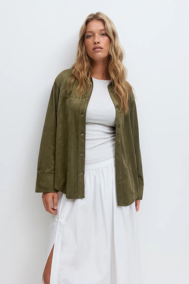 Surchemise boutonnée en daim vert olive portée sur un t-shirt blanc ajusté à col ras du cou, associée à une jupe longue blanche avec fente latérale.