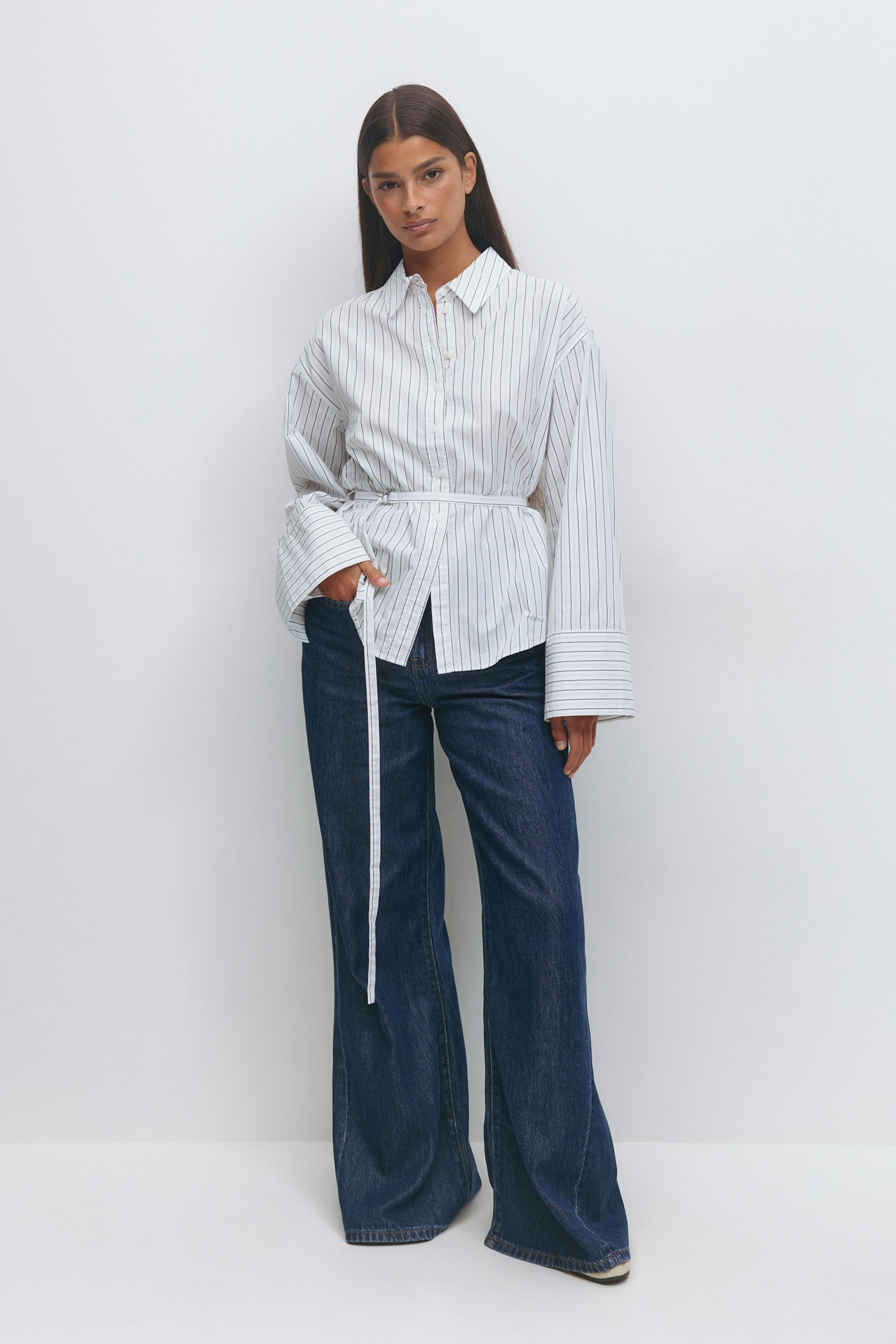 Hvid langærmet nålestribet skjorte med forlængede manchetter og et tyndt selvbinderbælte i taljen, sat sammen med mørke indigo jeans med brede ben.