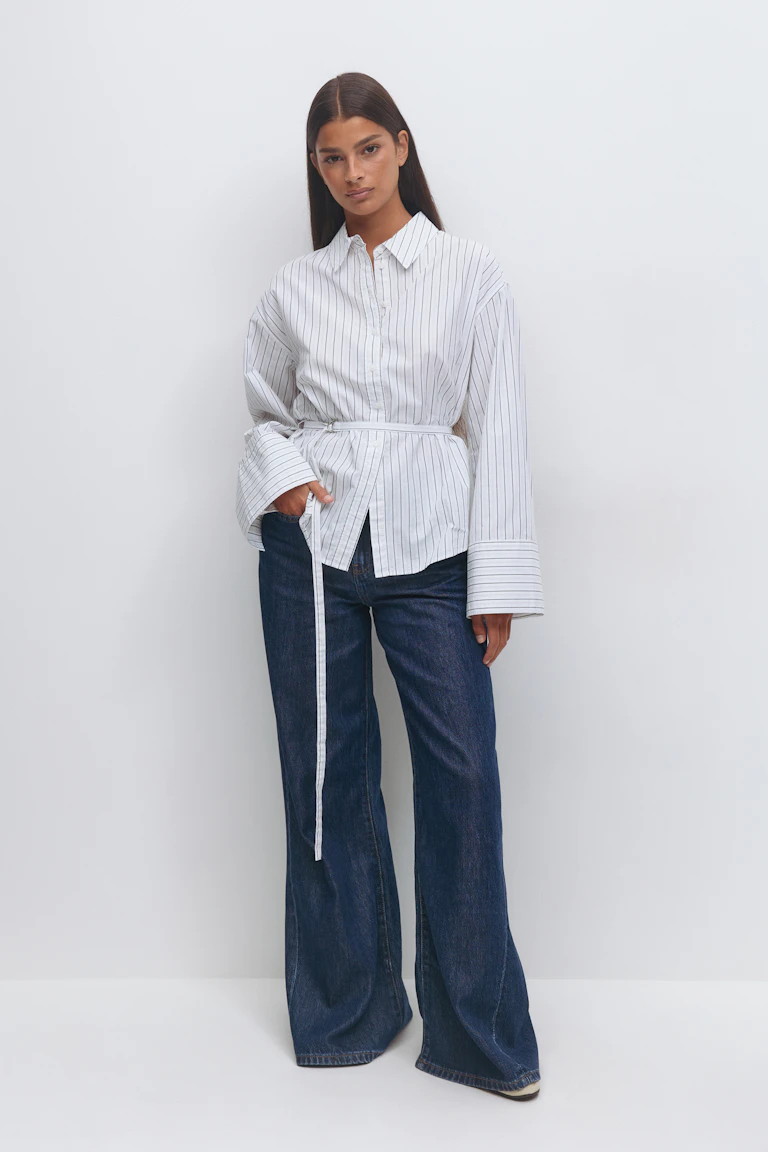 Hvit langermet, krittstripet skjorte med forlengede mansjetter og et tynt belte med selvknyting i midjen, sammen med mørke indigofargede jeans med vidde ben.