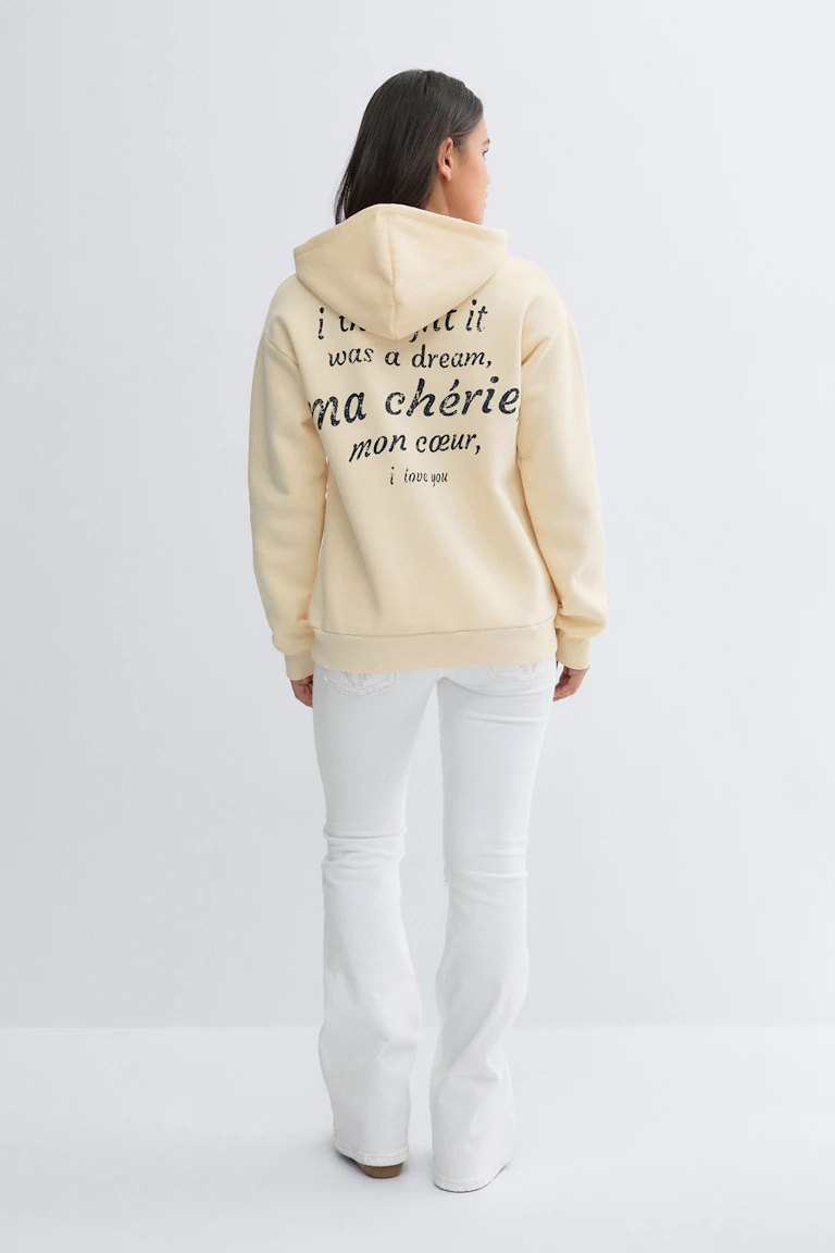 Pale yellow oversized hoodie with black printed script on the back, paired with white flared jeans and beige slip-on shoes.