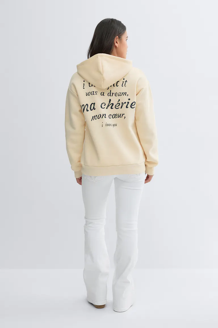 Lichtgele oversized hoodie met zwarte letters op de achterkant, gecombineerd met een witte flared jeans en beige slip-on schoenen.