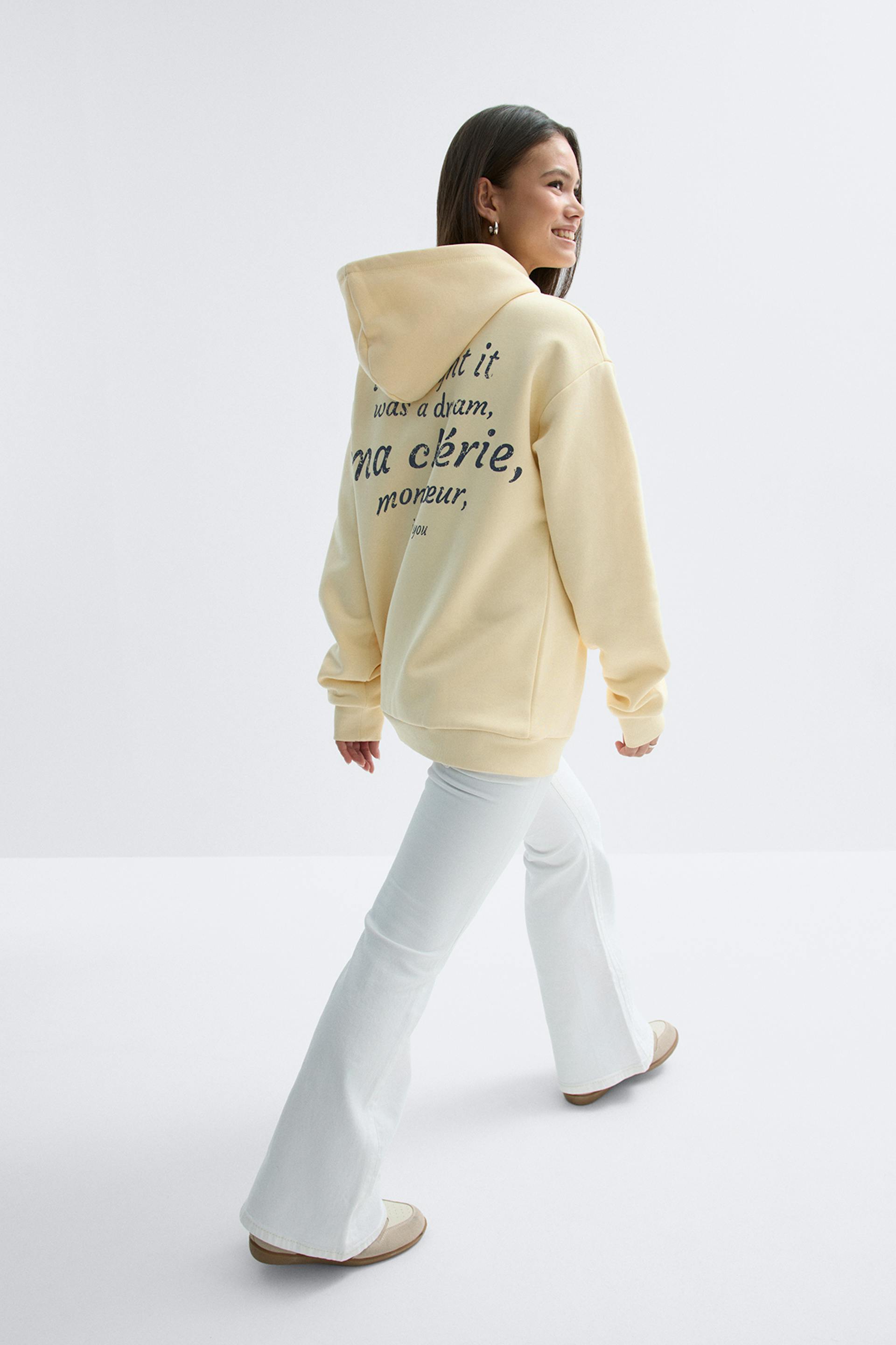 Sweat à capuche surdimensionné jaune pâle avec un imprimé noir au dos, associé à un jean évasé blanc et à des chaussures à enfiler beiges.