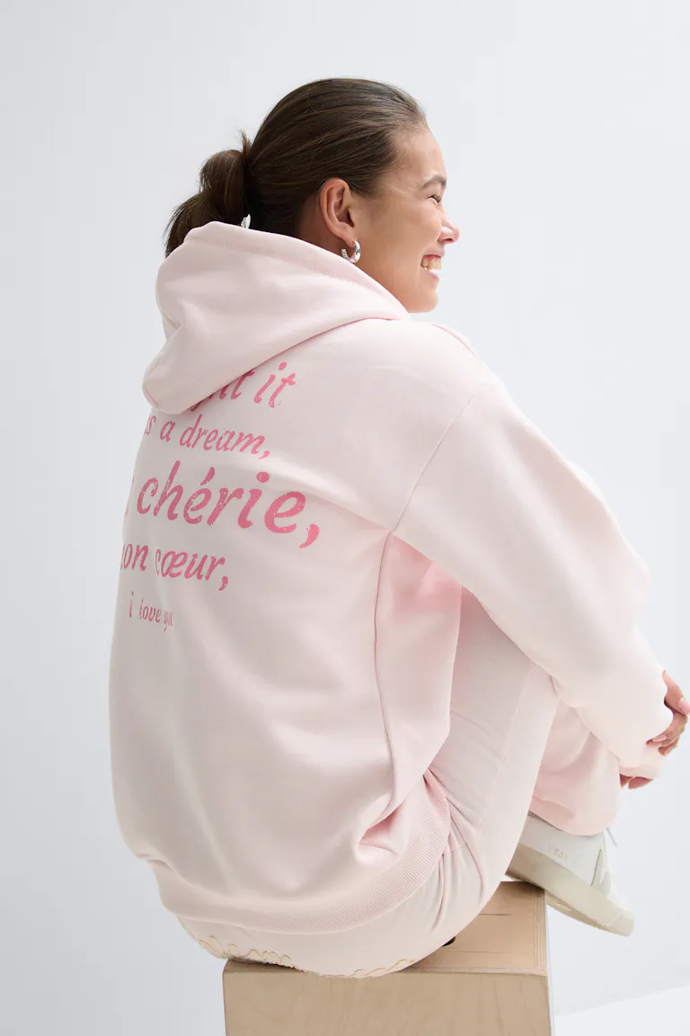 Pastel pink oversized hoodie with pink script on the back, paired with matching pink sweatpants and white low-top sneakers.