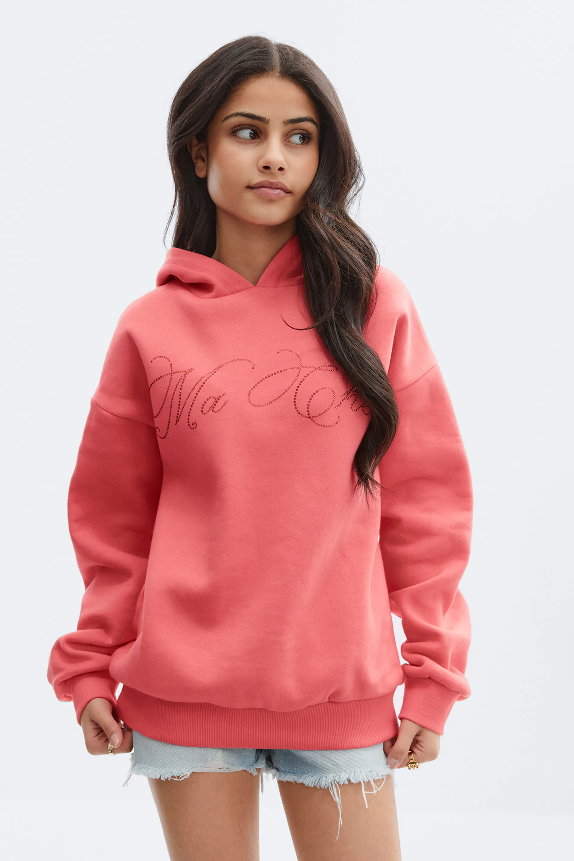 Korallenrotes Kapuzensweatshirt mit dezentem Strass-Schriftzug auf der Brust, kombiniert mit hellen, ausgefransten Denim-Shorts.