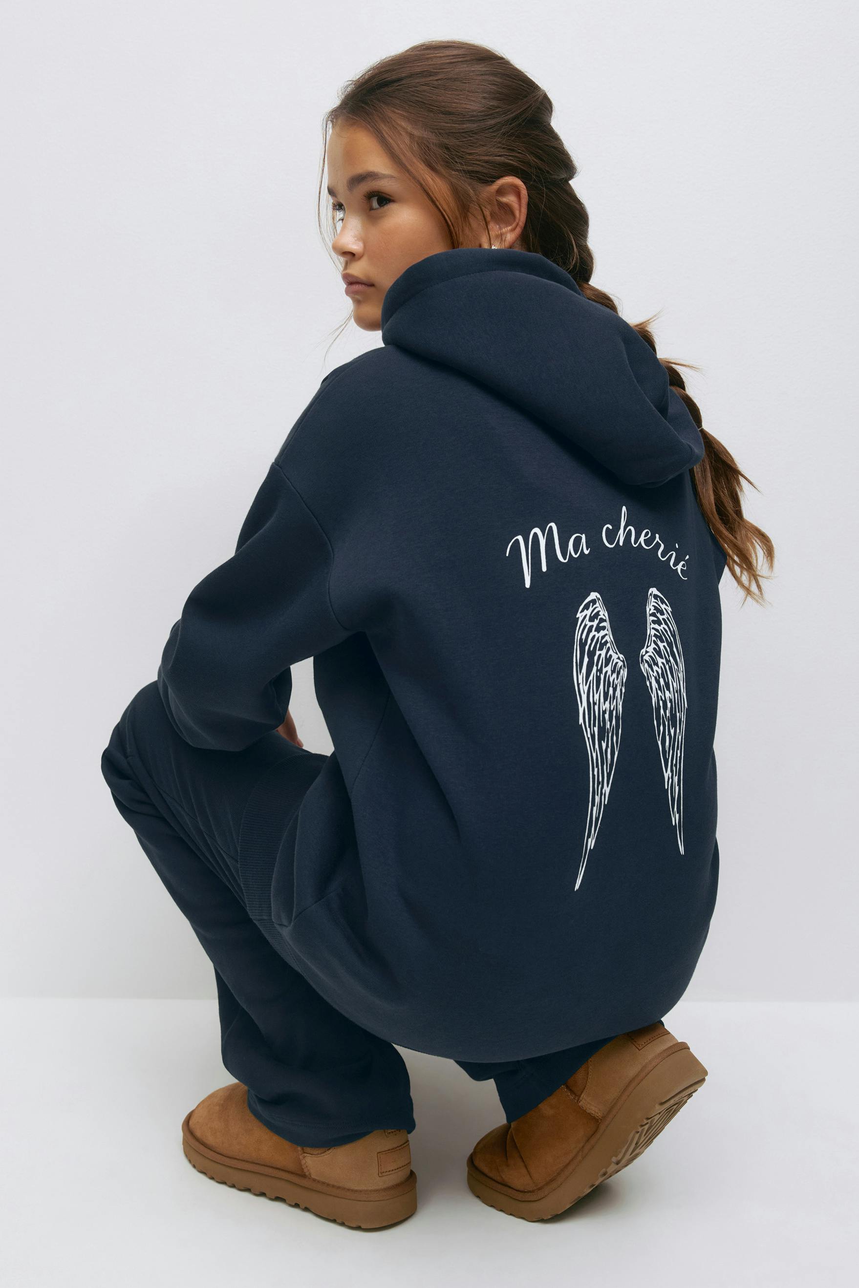 Navy oversized hooded sweatshirt featuring white "Ma chérie" script and angel wings print on the back, paired with matching navy sweatpants and tan suede slip‑on boots.